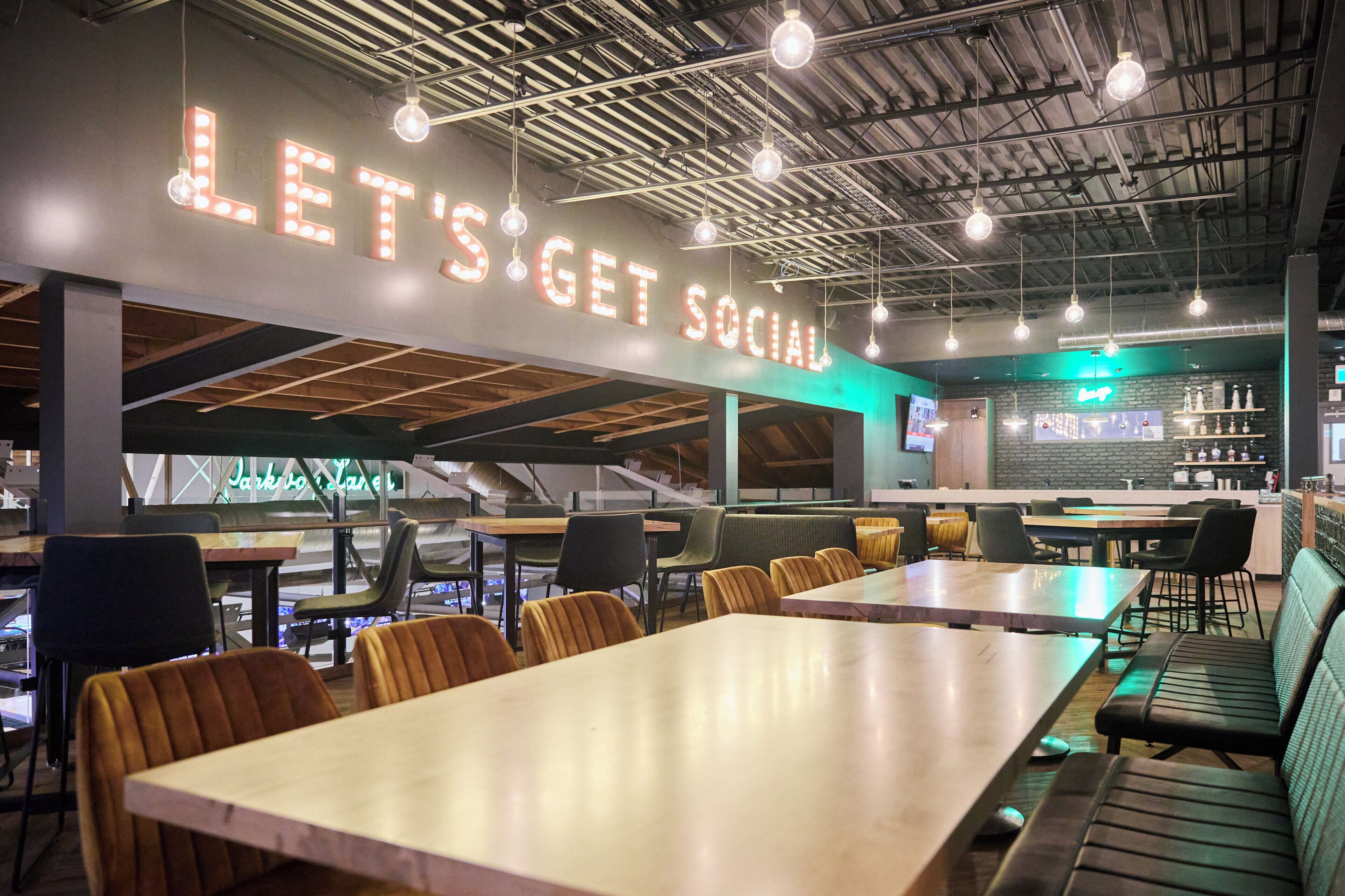 A modern cafe interior with a 'LET'S GET SOCIAL' sign, wooden tables, and green upholstered seating.