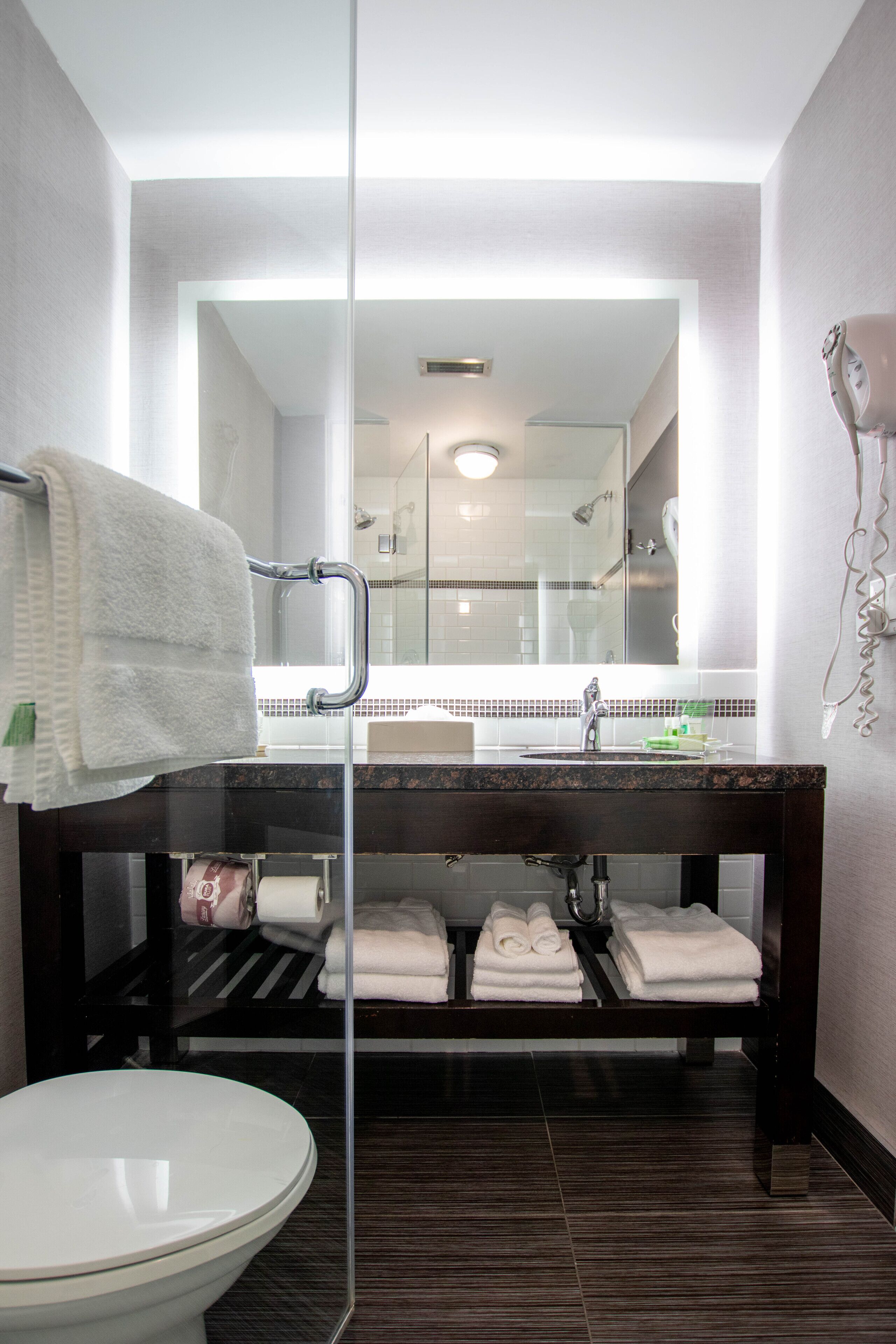 Un baño moderno con cabina de ducha de vidrio, inodoro blanco, tocador de madera oscura con espejo y toallas blancas dobladas con cuidado.
