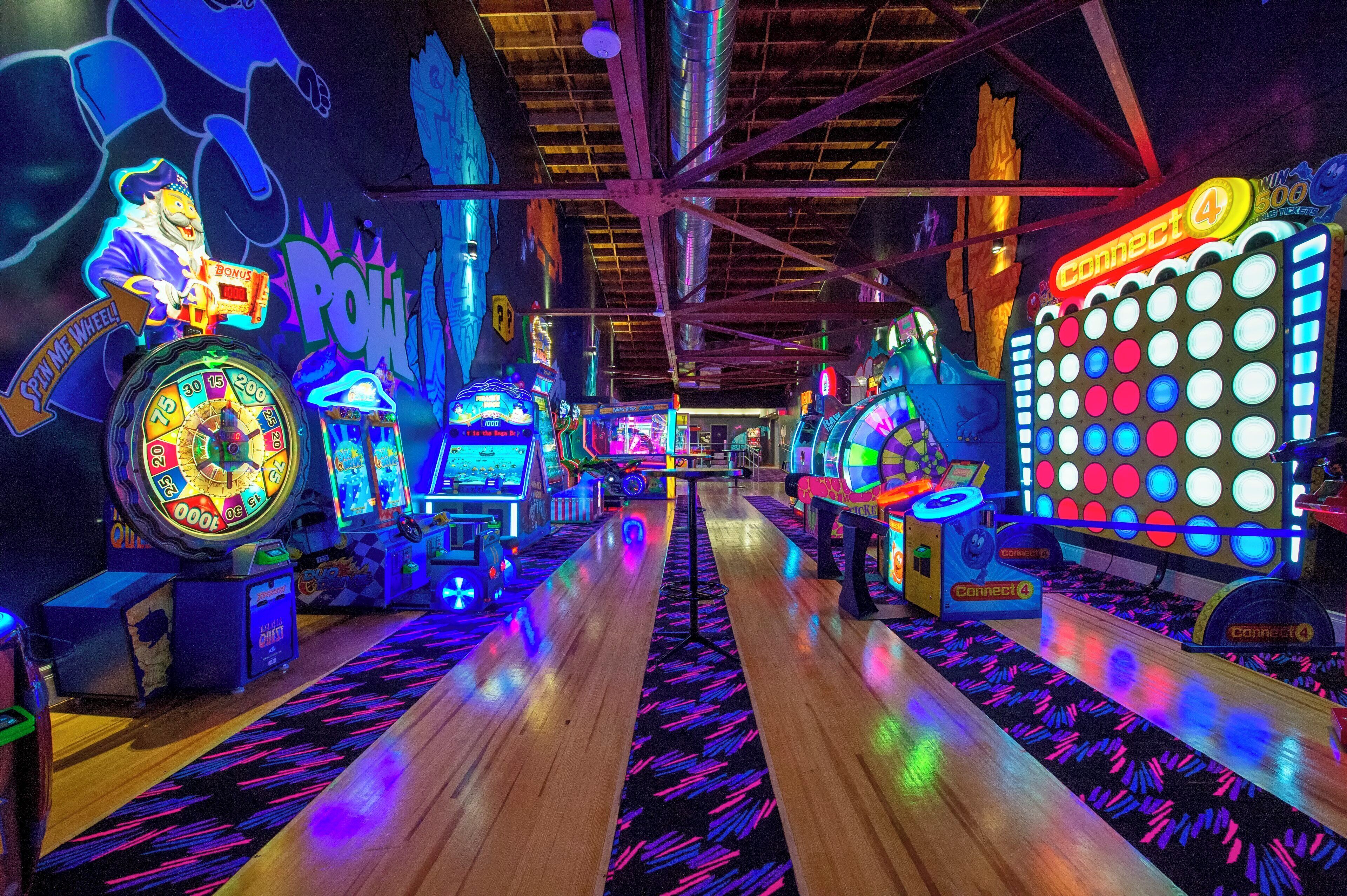 Un salón de juegos interior con varias máquinas recreativas, un pasillo central y un techo con vigas a la vista.