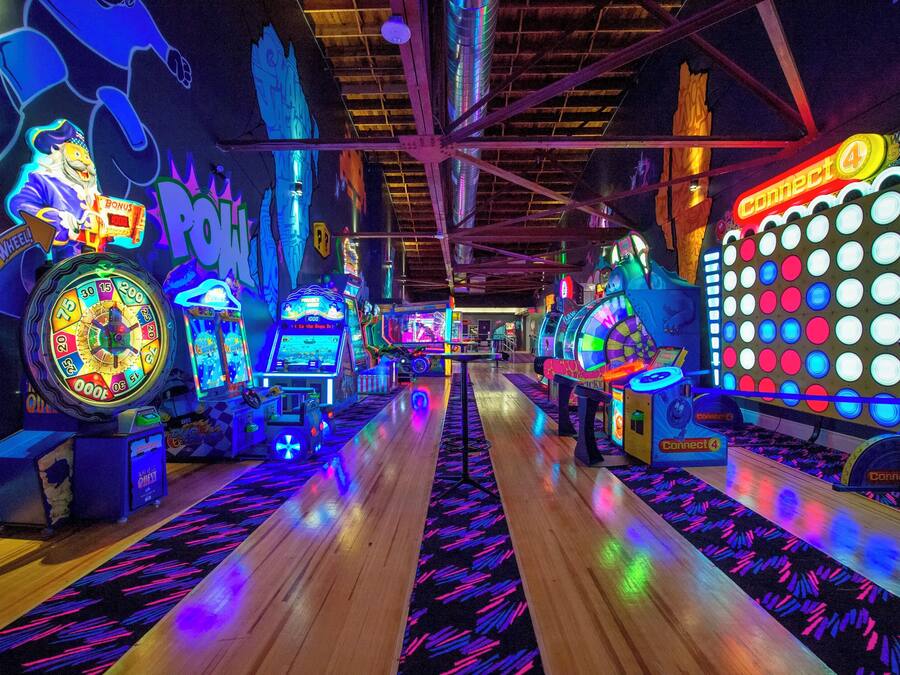 Un salón de juegos interior con varias máquinas recreativas, un pasillo central y un techo con vigas a la vista.