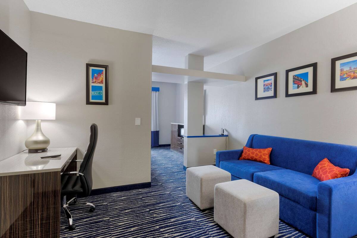 Habitación de hotel con escritorio, silla, sofá azul, otomanas e imágenes enmarcadas en la pared.