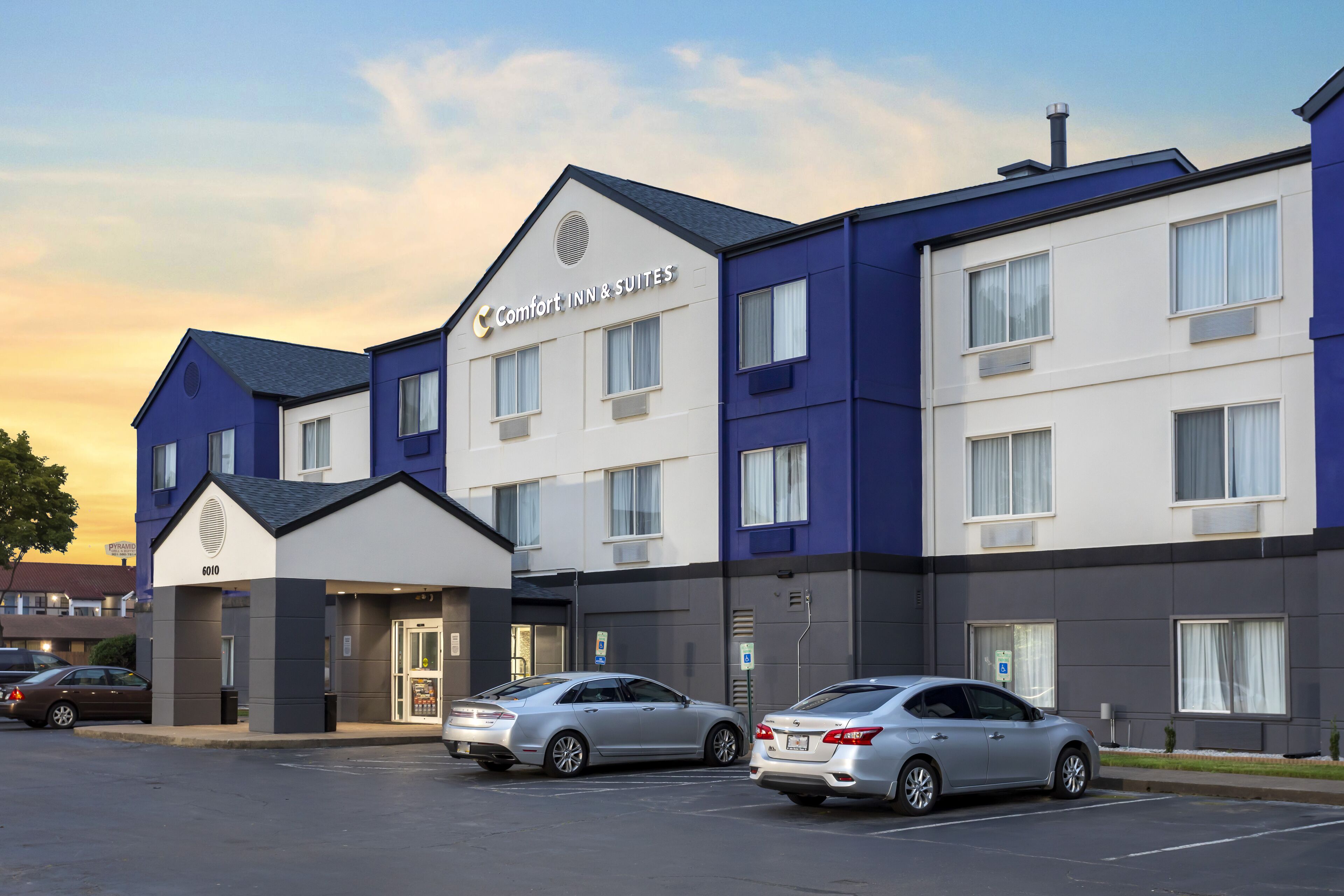 A modern hotel with a blue and white facade, two parking spaces, and a clear sky.