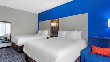 Habitación de hotel con dos camas, una pared azul decorativa, un espejo grande y un sofá azul con un cojín naranja.