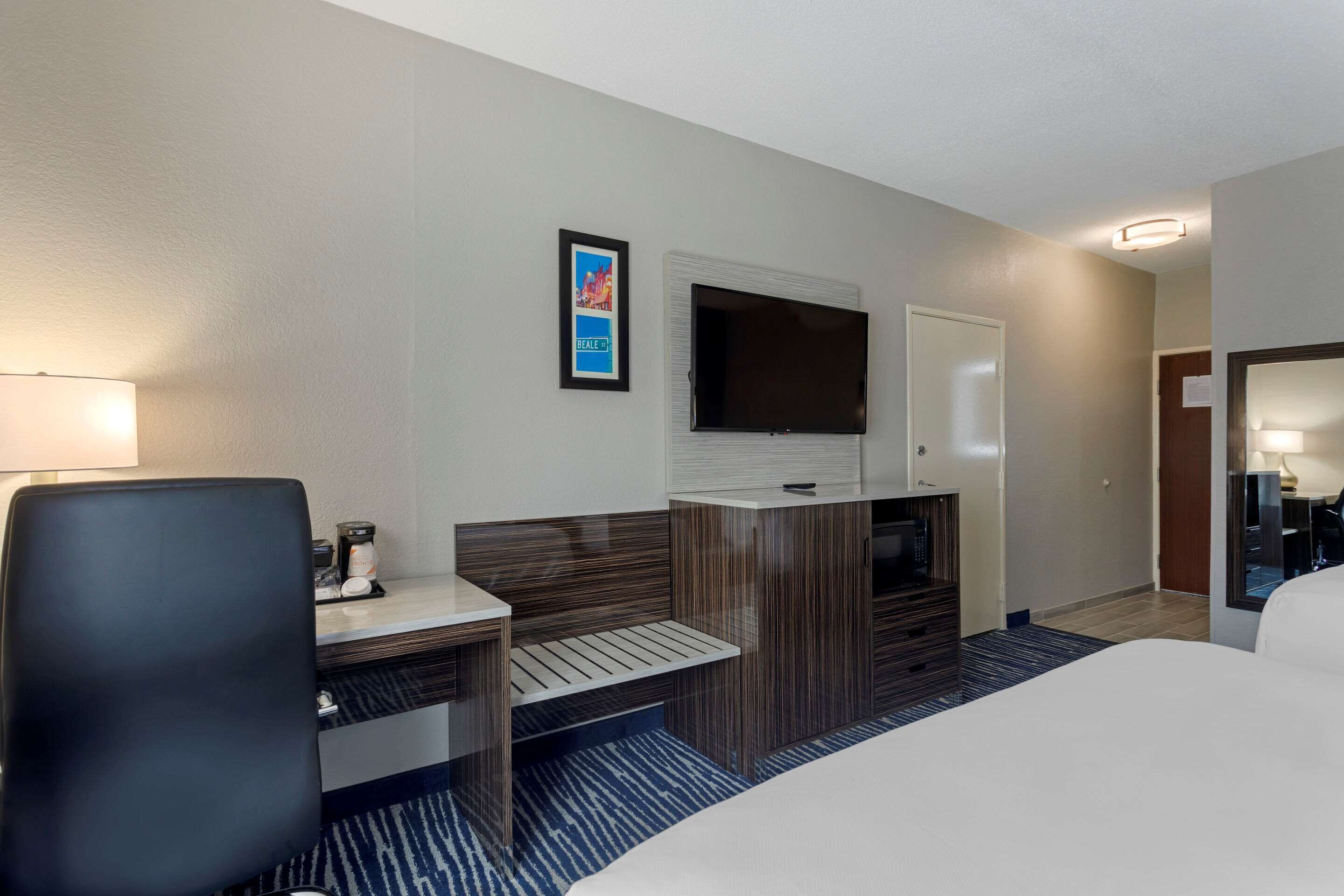 A hotel room with a bed, a desk with a chair, a TV mounted on the wall, and a small seating area with a bench.