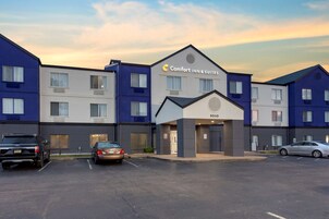 Un hotel con diseño moderno, destacando un letrero grande de 'Comfort Inn & Suites'. Tiene varios pisos con diferentes estilos de ventanas y un estacionamiento con varios coches.