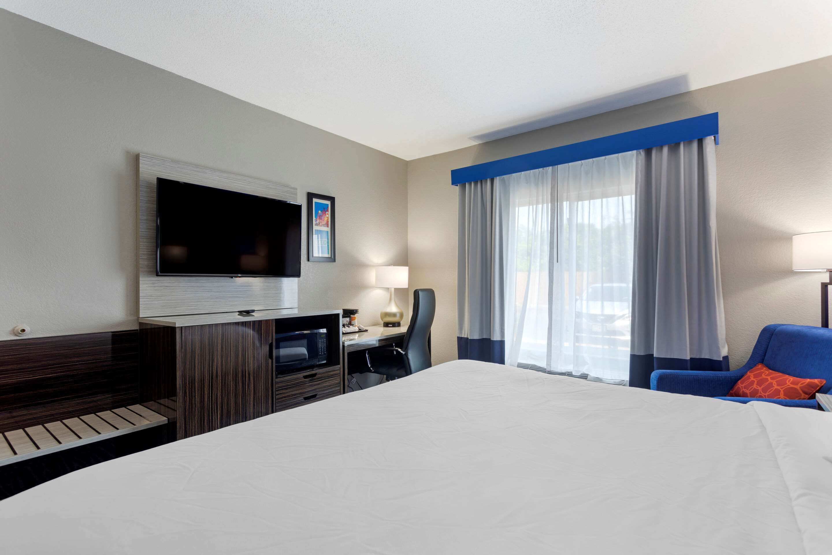 Una habitación de hotel con una cama, un escritorio con computadora, un televisor montado en la pared y una ventana con cortinas.