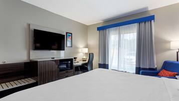 Una habitación de hotel con una cama, un escritorio con computadora, un televisor montado en la pared y una ventana con cortinas.