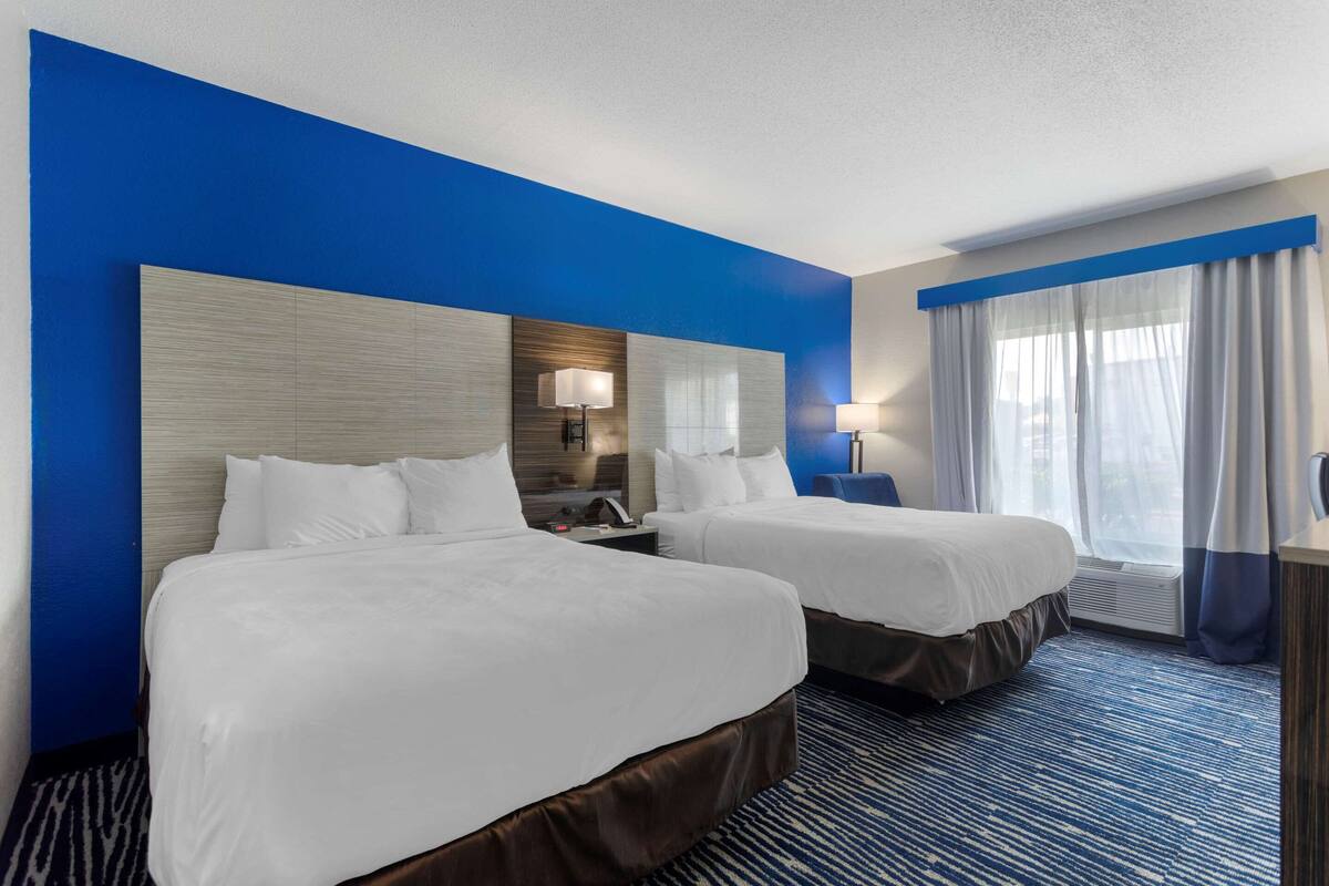 Habitación de hotel con dos camas, un espejo grande, un escritorio con lámpara y una ventana con cortinas.