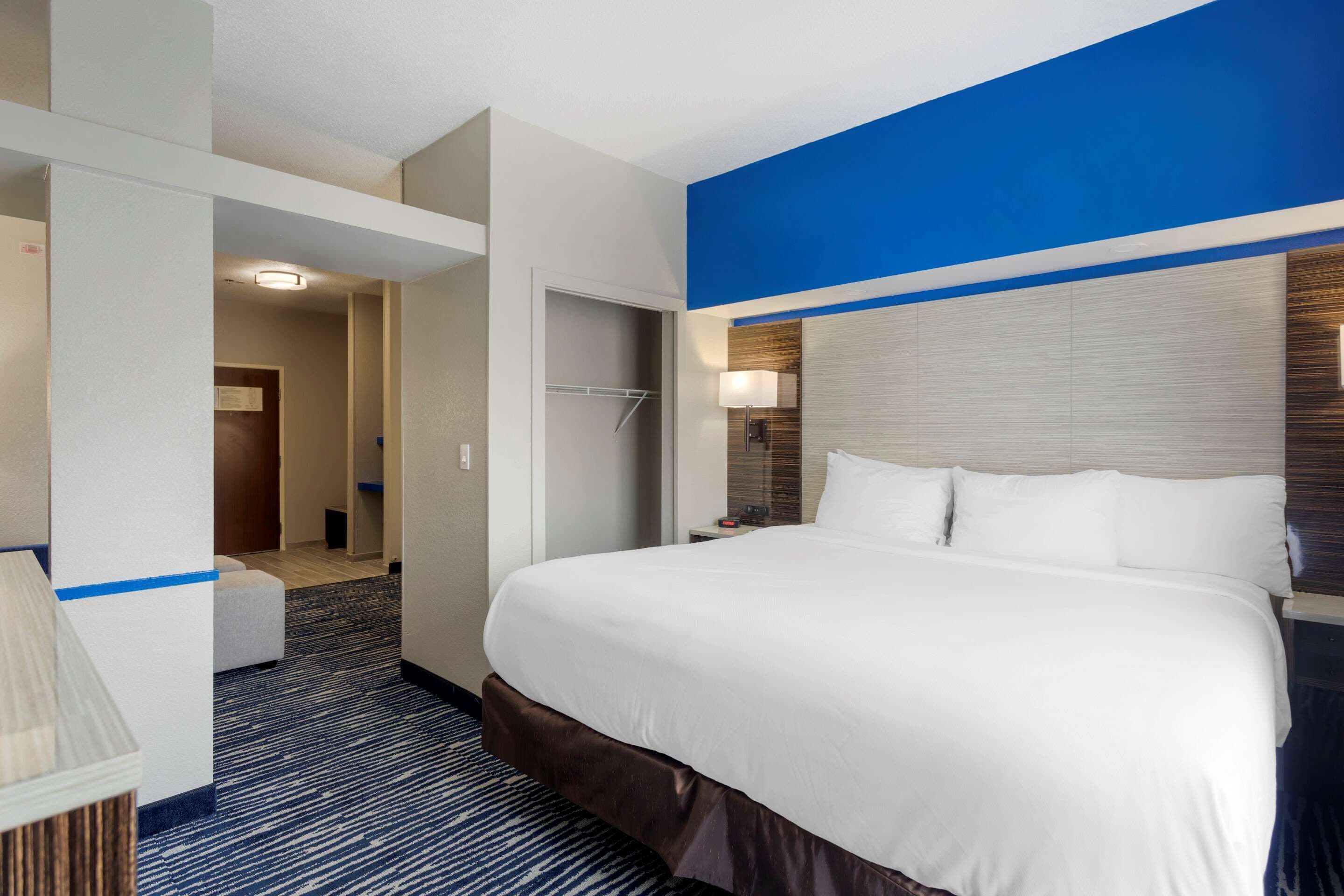 A hotel room with a large bed, a nightstand, a lamp, a wardrobe, and a door leading to another room.