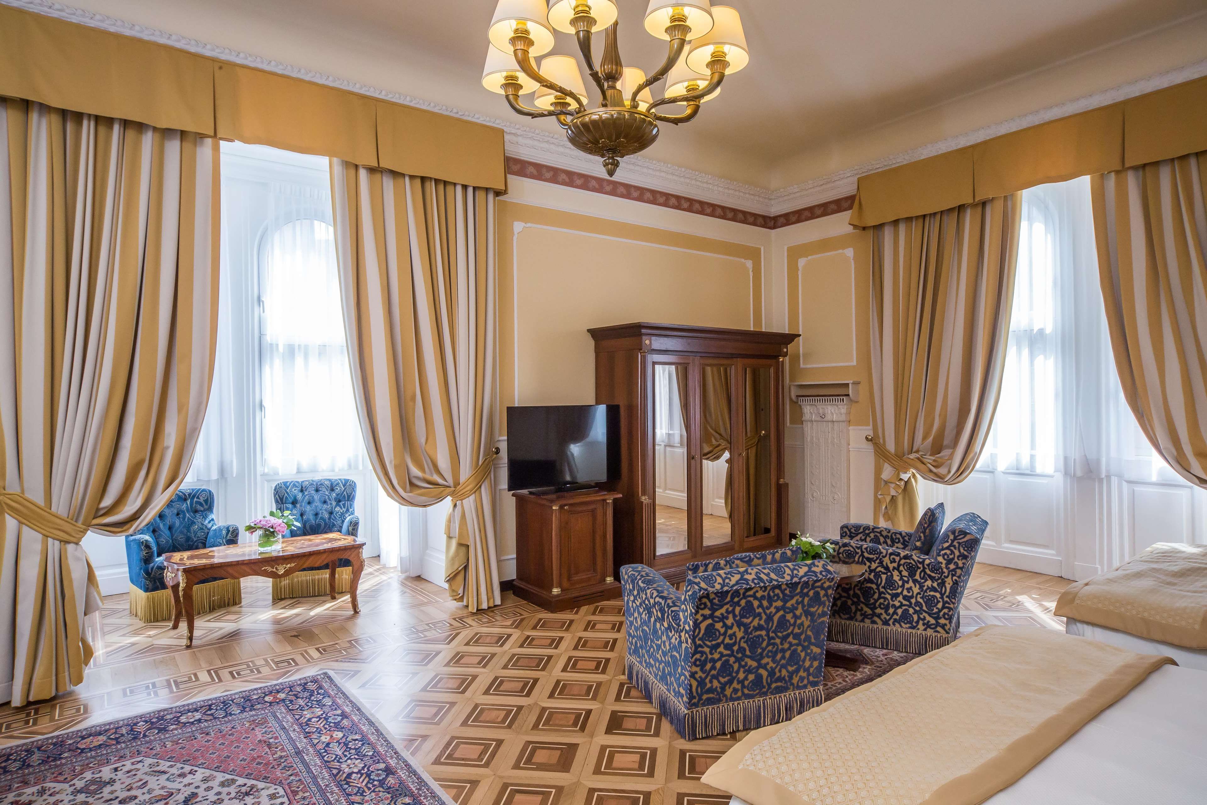 Habitación de hotel con una cama grande, televisión, aparador de madera, dos sillones estampados, una mesita pequeña y una alfombra decorativa.