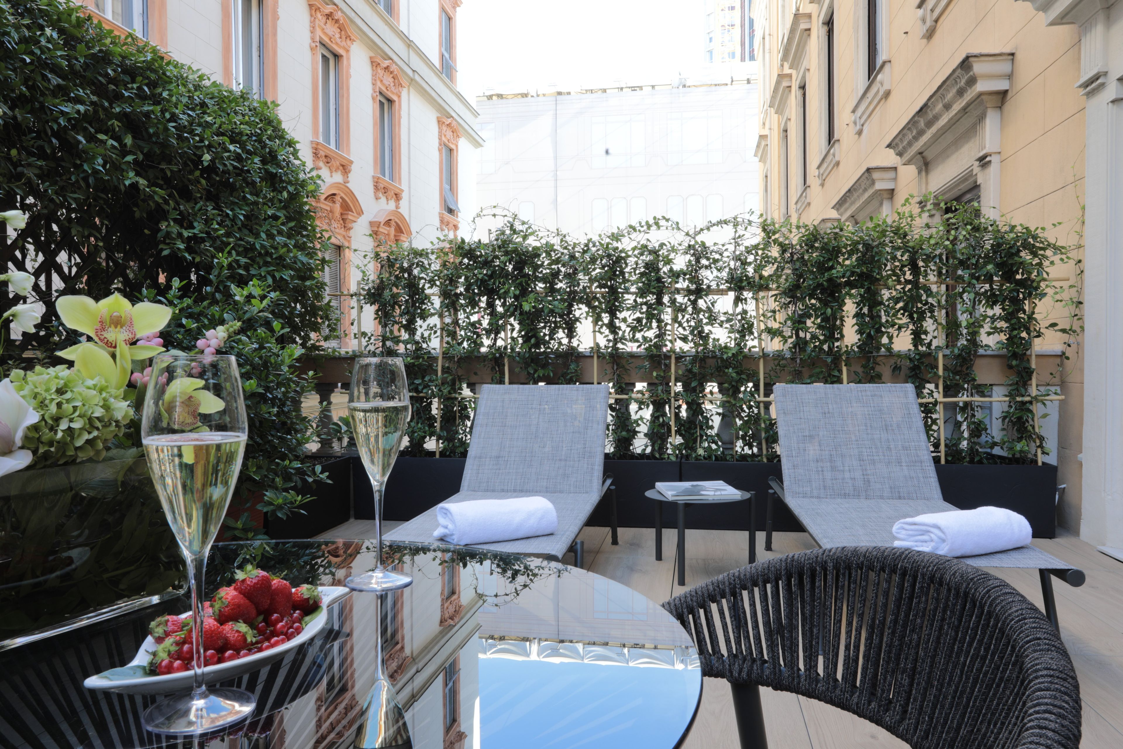 A terrace with two lounge chairs, a table with a glass of wine and a bowl of strawberries, and a backdrop of greenery and buildings.