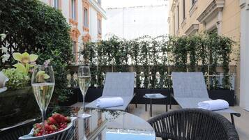 A terrace with two lounge chairs, a table with a glass of wine and a bowl of strawberries, and a backdrop of greenery and buildings.