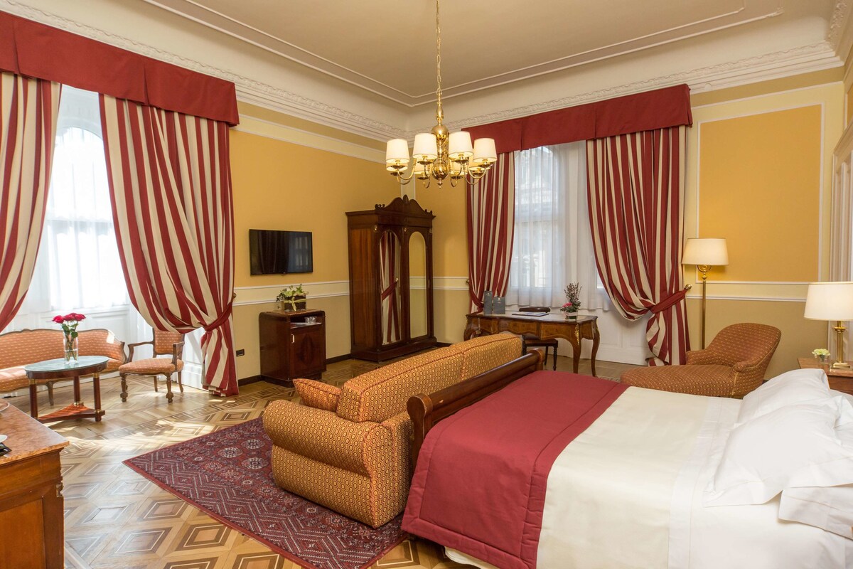 Habitación de hotel con una cama grande, dos sillones, un sofá, una mesita de madera, un espejo y una araña.