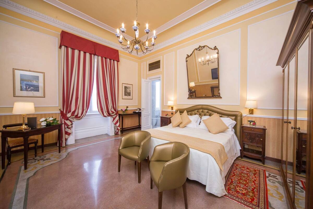 A hotel room with a large bed, two chairs, a desk, a chandelier, a mirror, and a red and white striped curtain.