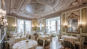 Un comedor formal con mesas redondas, sillas blancas, candelabros y detalles ornamentales en el techo.