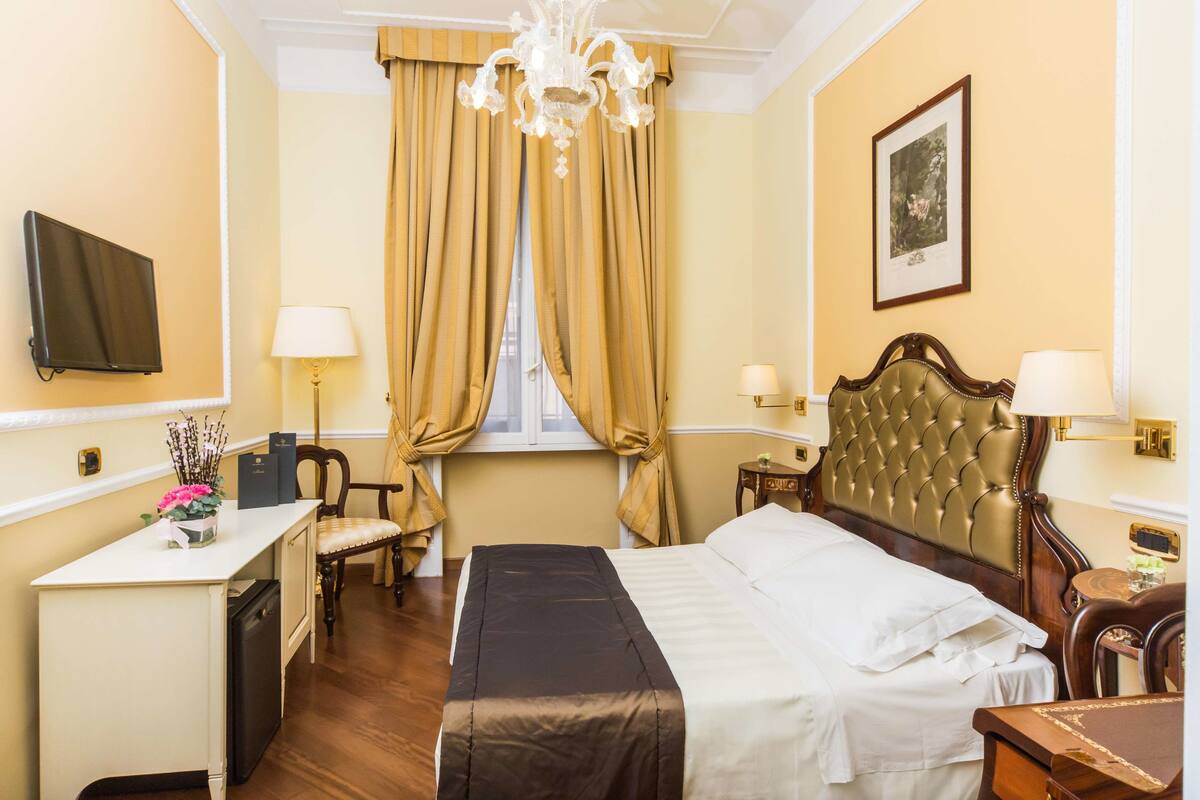 A hotel room with a bed, bedside tables, a desk, a chair, a television, a chandelier, and curtains.