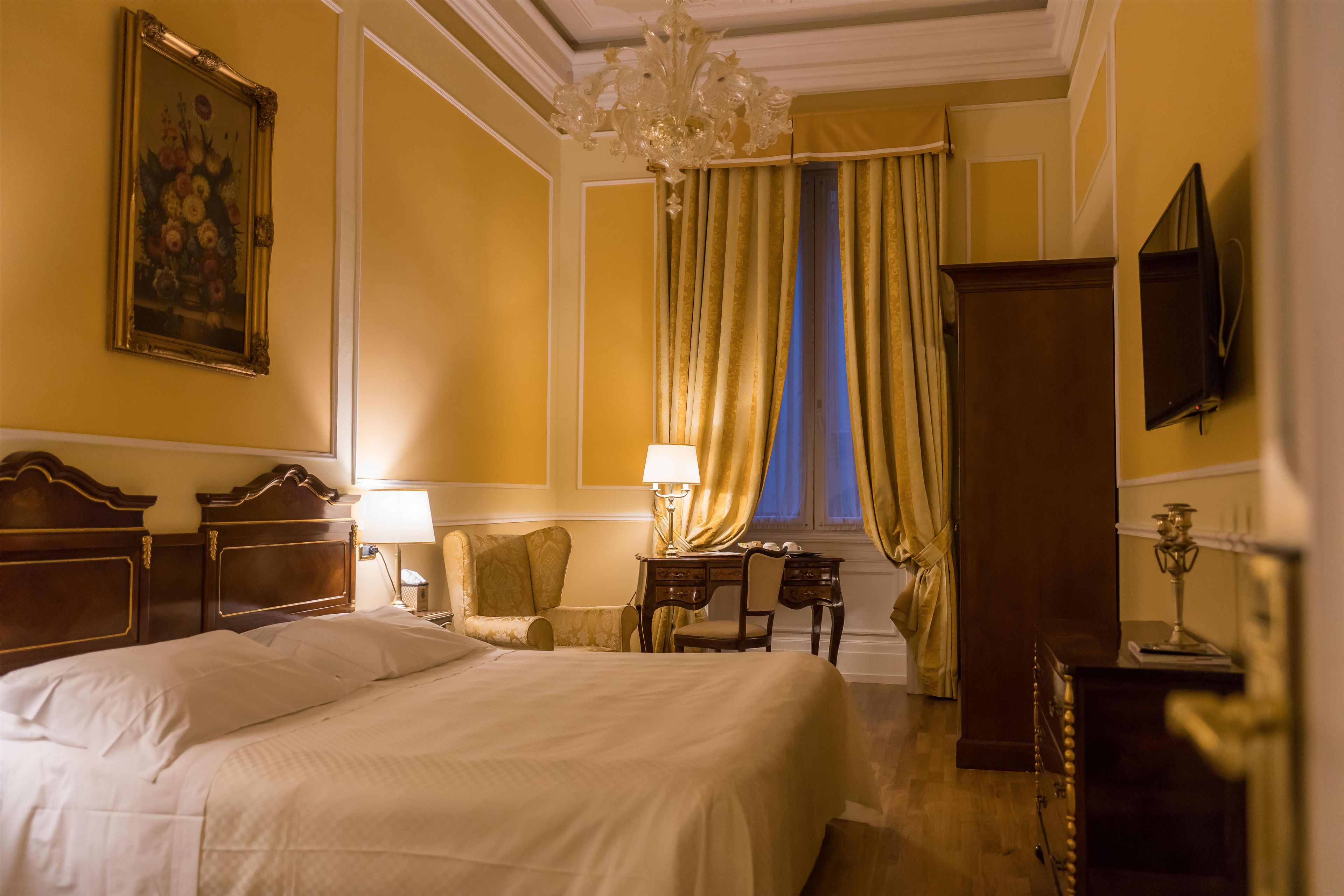 A hotel room with a large bed, a desk with a lamp, a chair, a television, and a window with curtains.