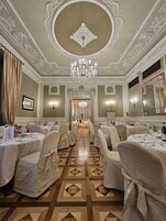 A formal dining room with ornate ceiling and chandeliers, arranged tables with chairs, and a patterned floor.