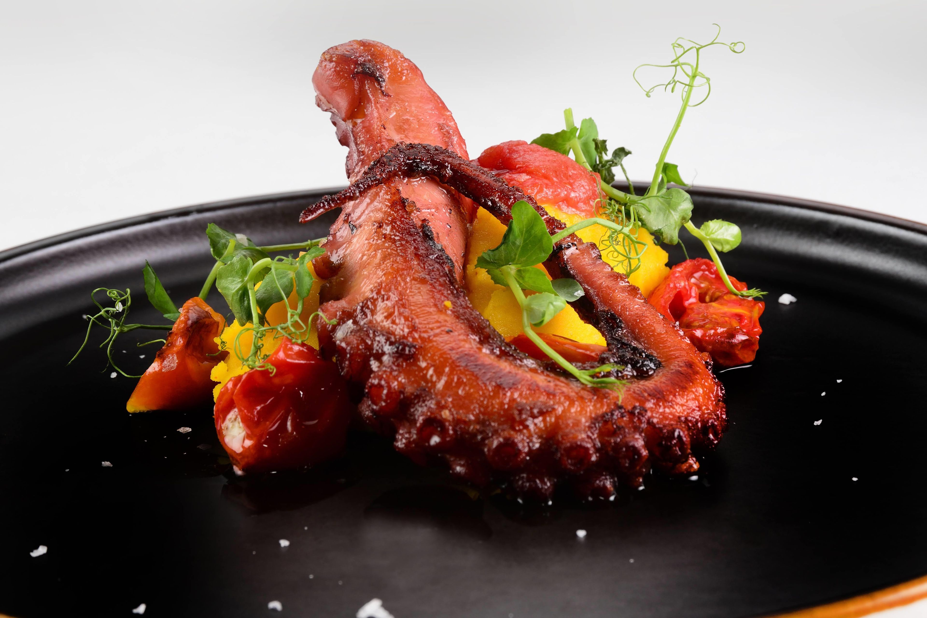 Un plat composé de poulpe rôti, de légumes et de verdure, servi dans une assiette noire.