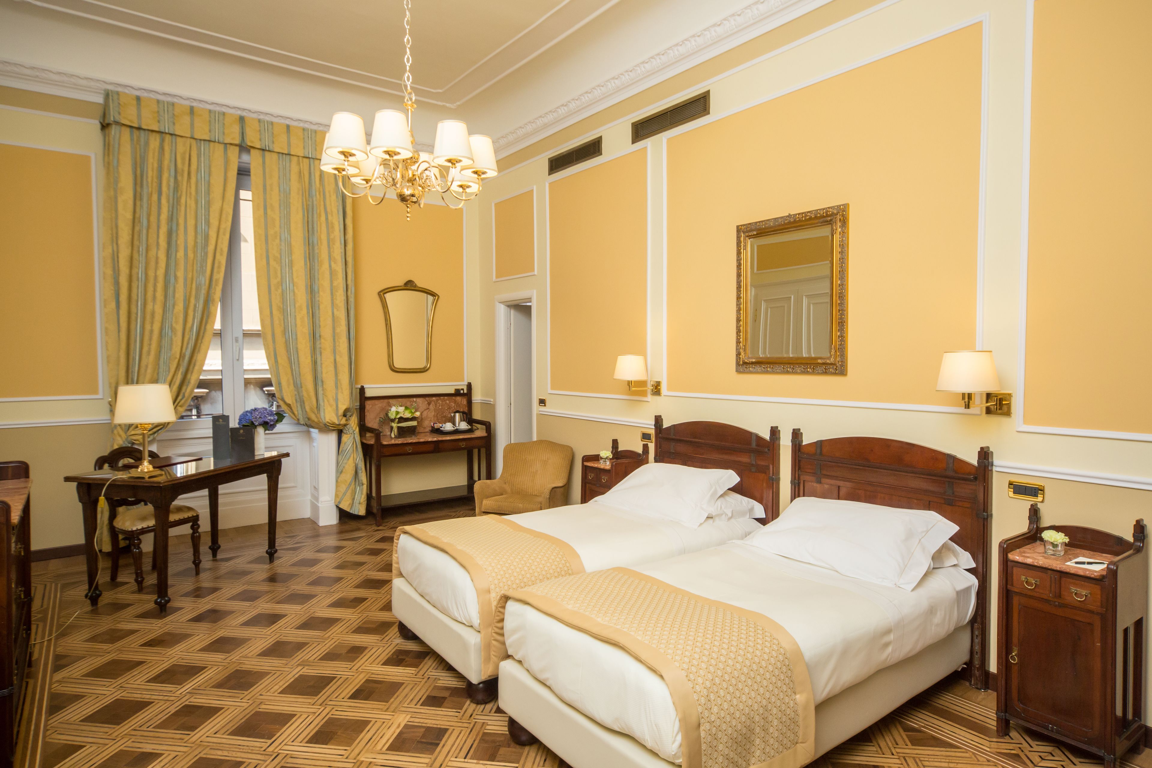 A hotel room with two beds, a desk, a chair, a mirror, and a chandelier.