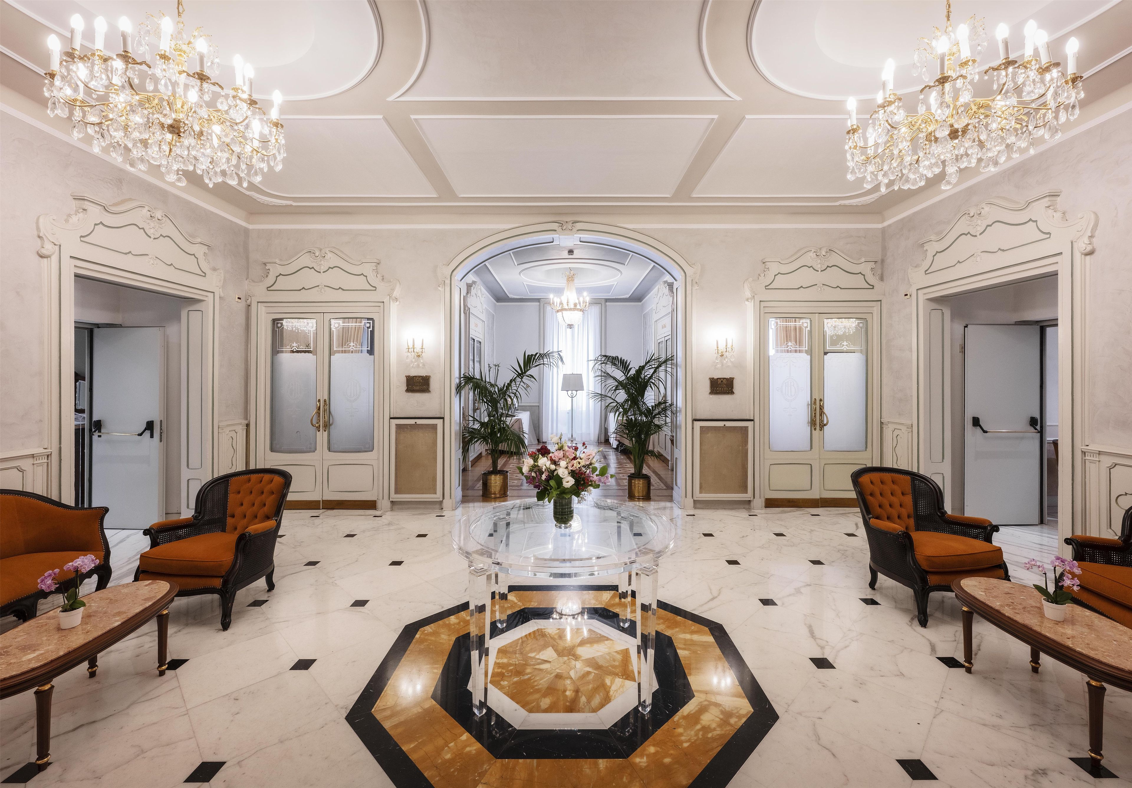 A grand lobby with a central glass table, ornate chandeliers, and classic furniture.