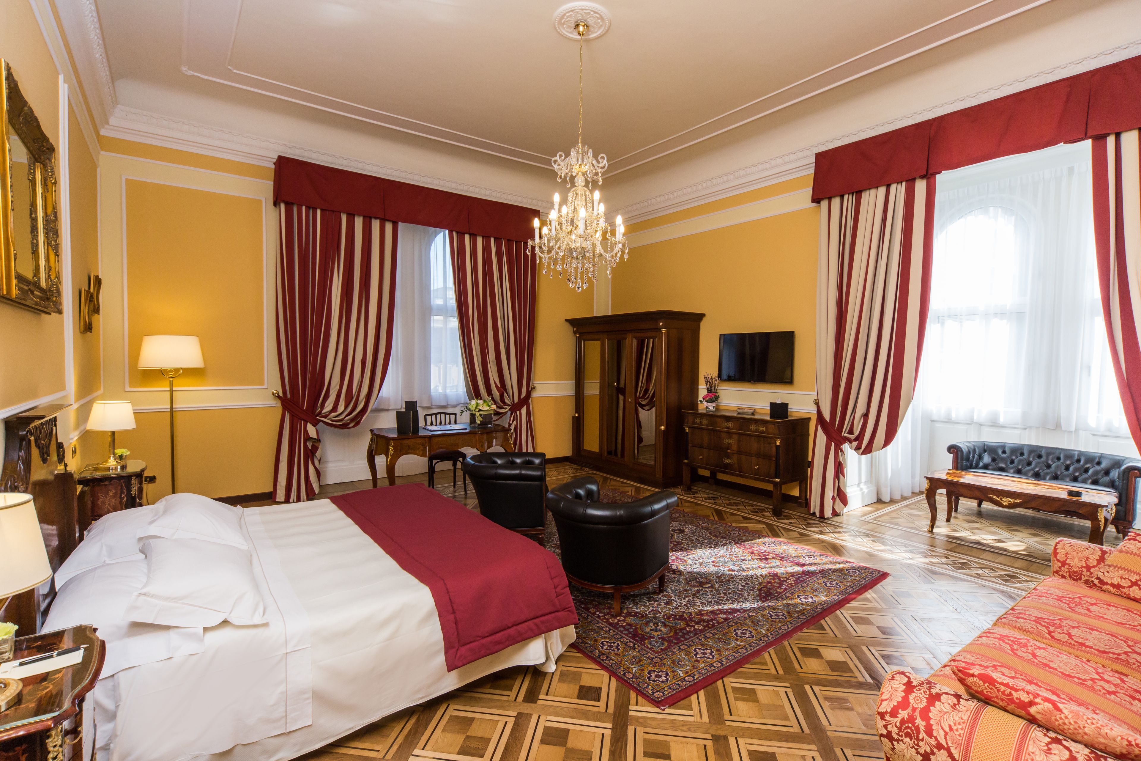 A hotel room with a large bed, a sitting area with a sofa and armchairs, a wooden floor, and a chandelier.