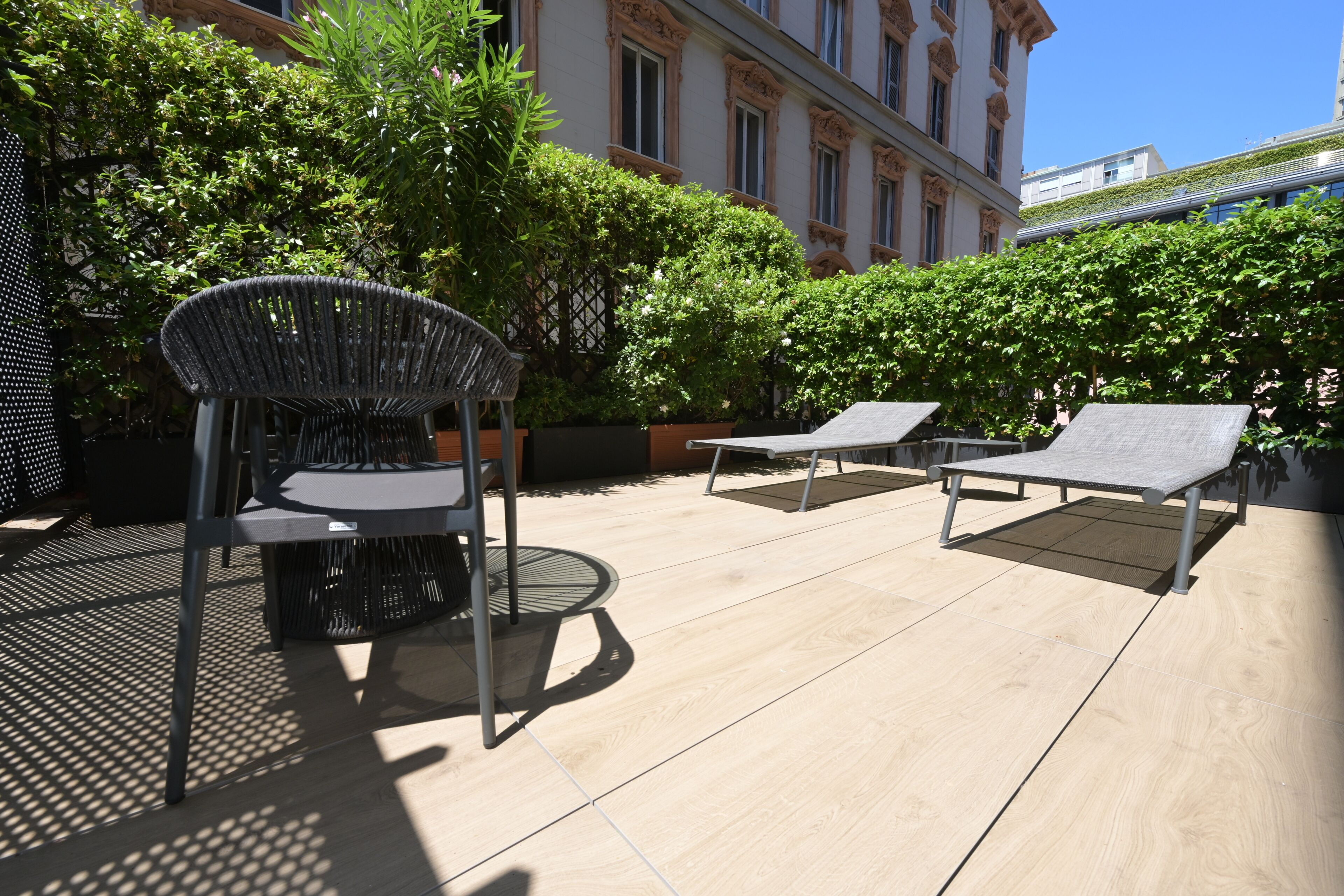 Un espace extérieur aménagé avec des chaises et des tables en métal, entouré de verdure et avec un bâtiment en arrière-plan.