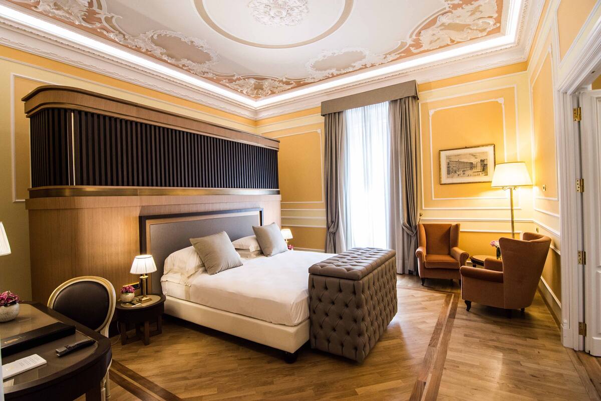 A hotel room with a large bed, a sitting area with a chair and a small table, a desk with a lamp, and a ceiling with decorative molding.