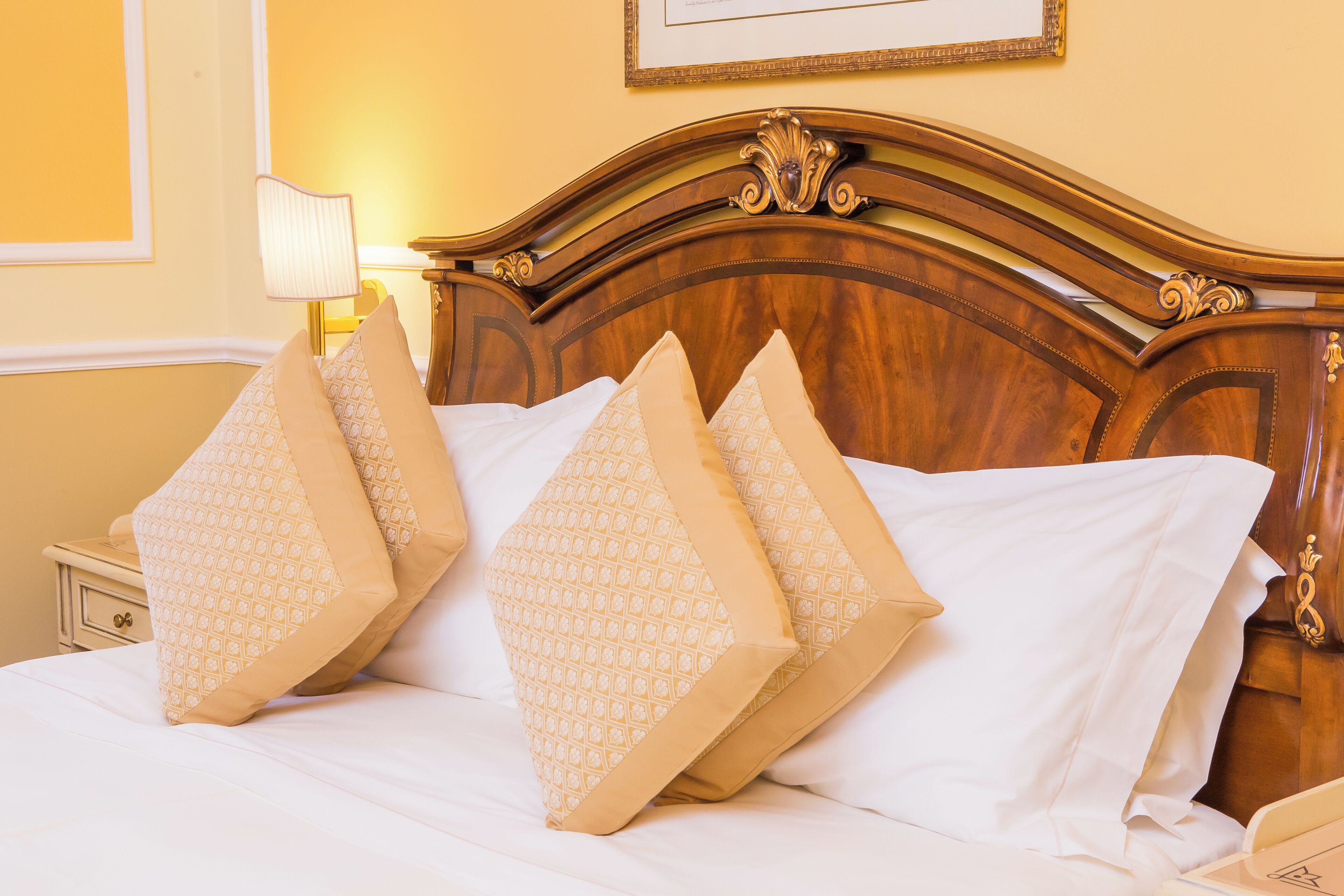 Een bed met vier gekleurde kussens, een houten hoofdbord, een nachtkastje met een lamp en een ingelijste foto aan de muur.