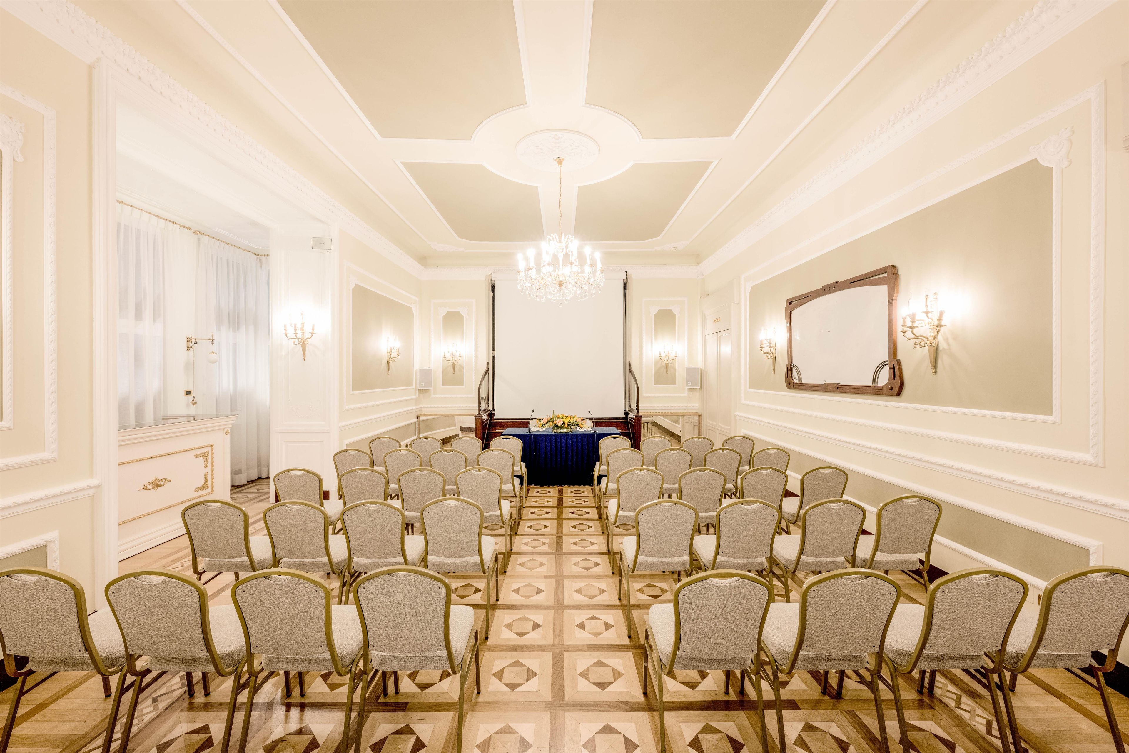A conference room with a long table, rows of chairs, a chandelier, and wall sconces.