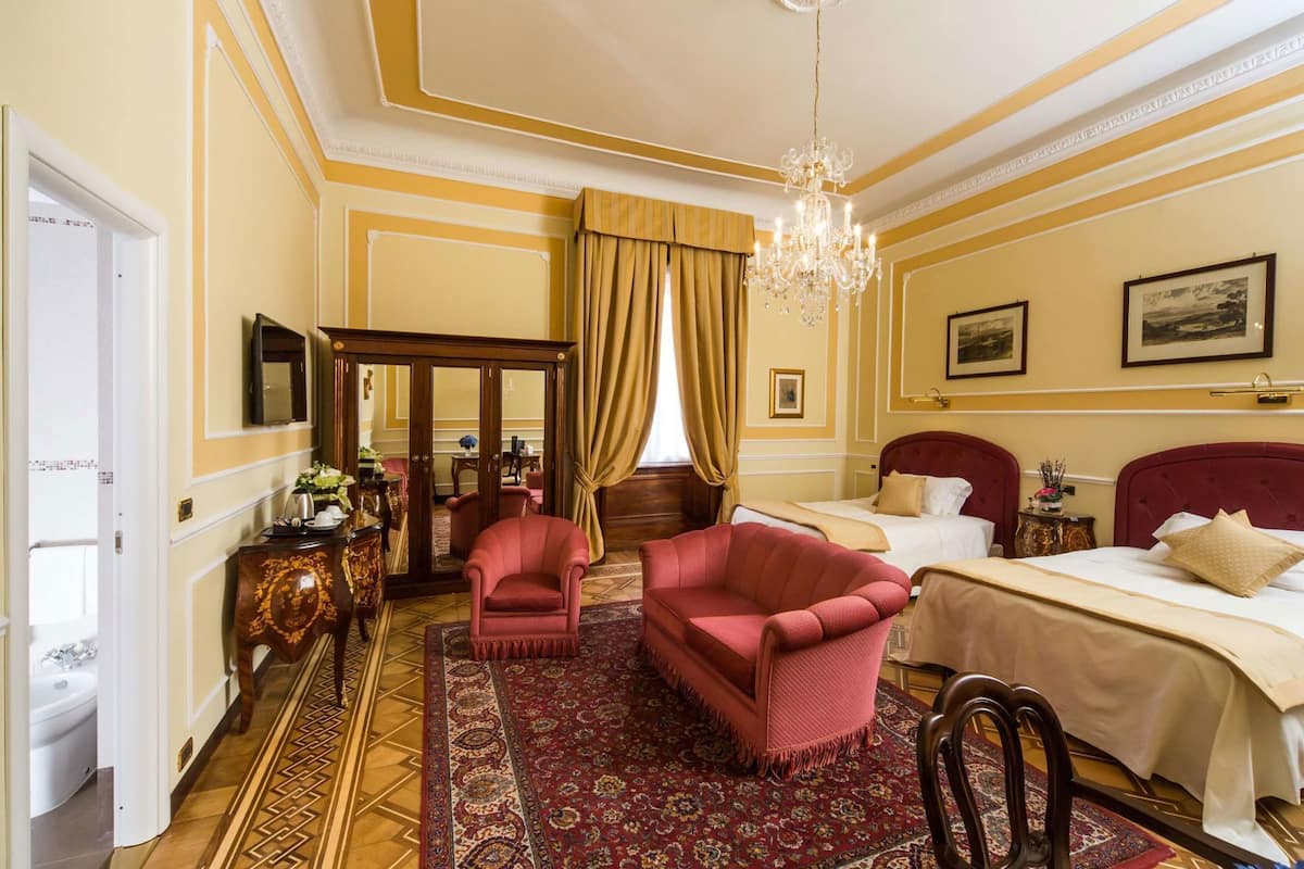 A hotel room with a large bed, two armchairs, a wooden wardrobe, a chandelier, and a patterned rug.