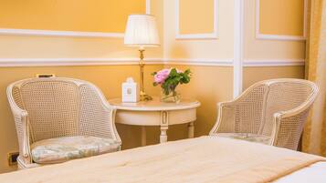 Una camera d'albergo con un letto, due poltrone in vimini, un tavolino rotondo con una lampada e fiori, e una TV a parete.