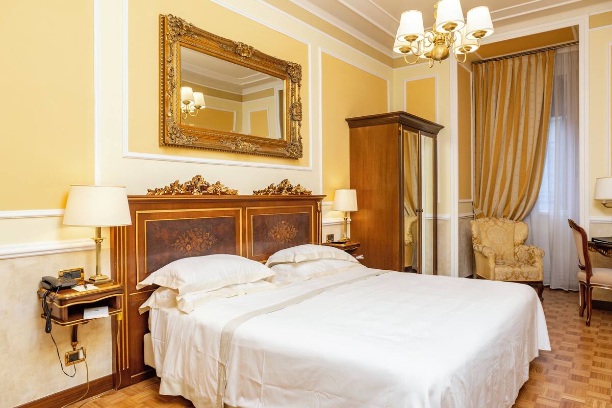 A hotel room with a large bed, two bedside tables with lamps, a wooden wardrobe, a chair, and a large mirror on the wall.