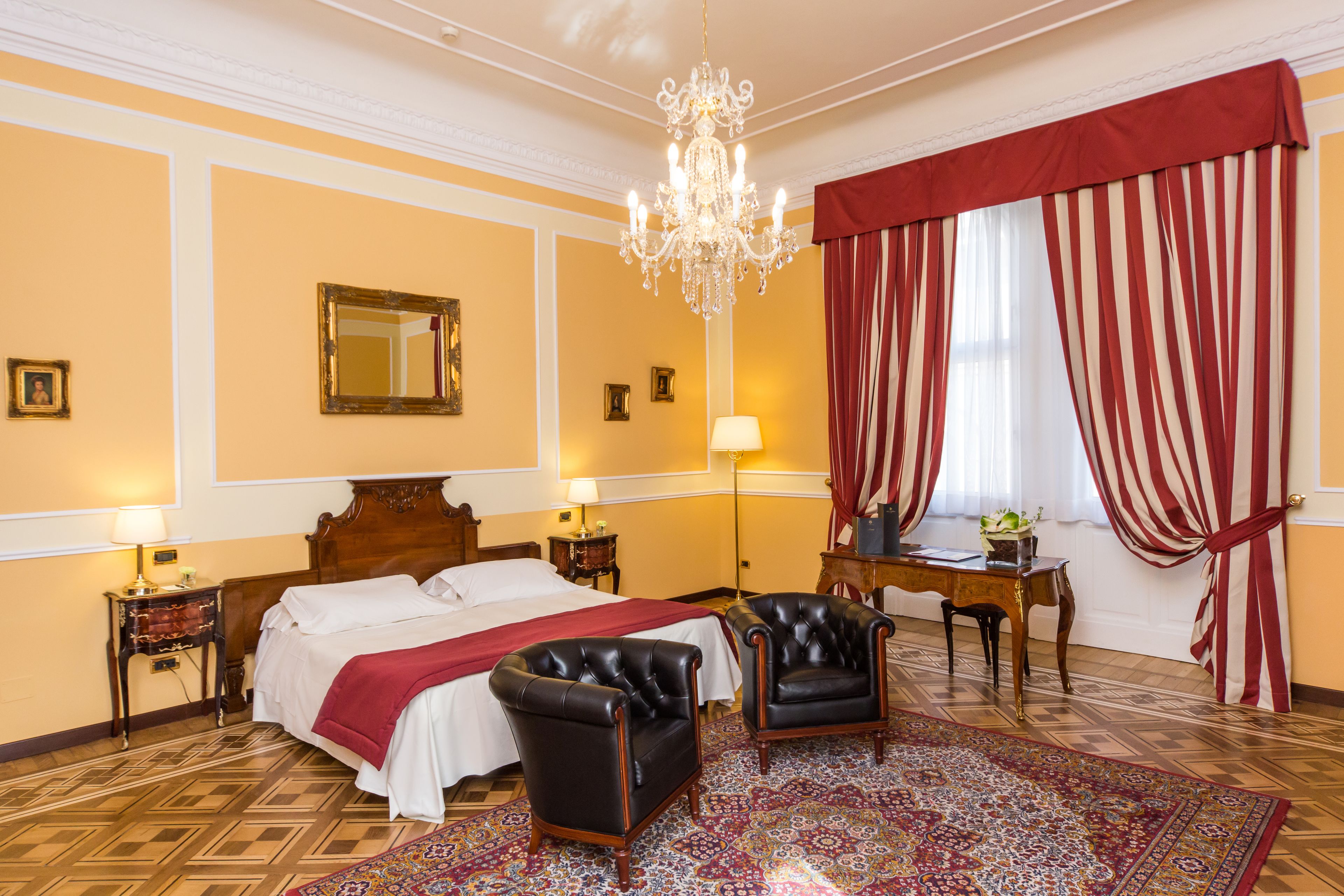 A hotel room with a large bed, two armchairs, a wooden desk, a chandelier, and a patterned rug.