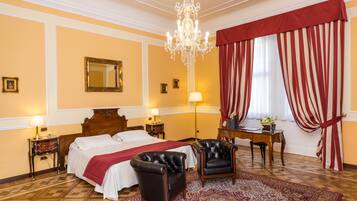 A hotel room with a large bed, two armchairs, a wooden desk, a chandelier, and a patterned rug.