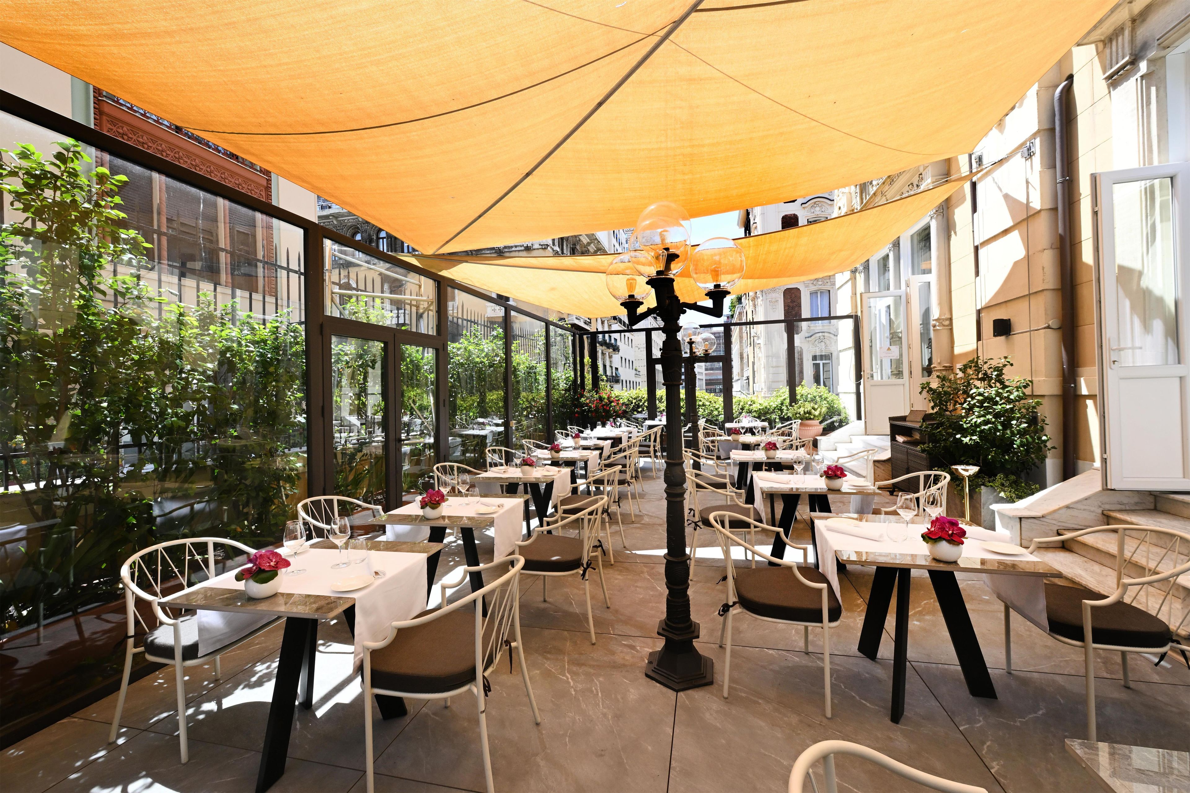 Zona pranzo all'aperto con tavoli e sedie, ampie finestre e vista sulla strada.