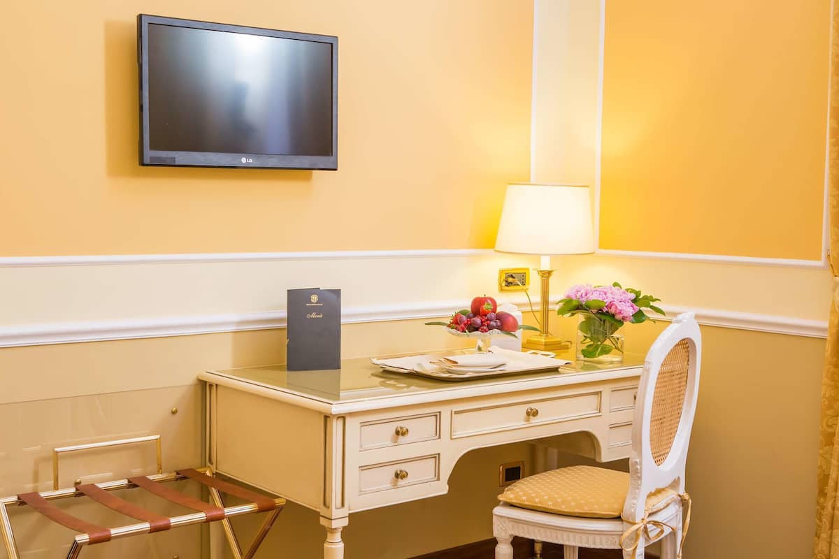 A hotel room with a desk, a television, a lamp, a chair, and a small table with a vase of flowers.