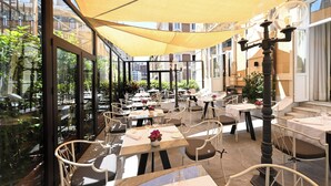 Un área de comedor al aire libre con mesas y sillas, ventanas grandes y un dosel amarillo.