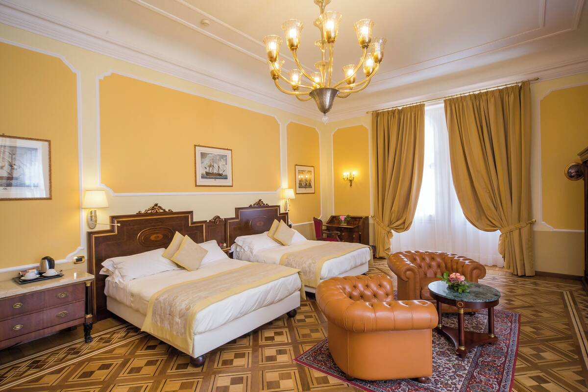 A hotel room with two beds, a chandelier, a leather armchair, and a small round table.