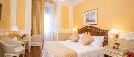 Une chambre d’hôtel avec un grand lit, deux tables de chevet avec des lampes, une tête de lit en bois, une chaise et une table de nuit avec des fruits.