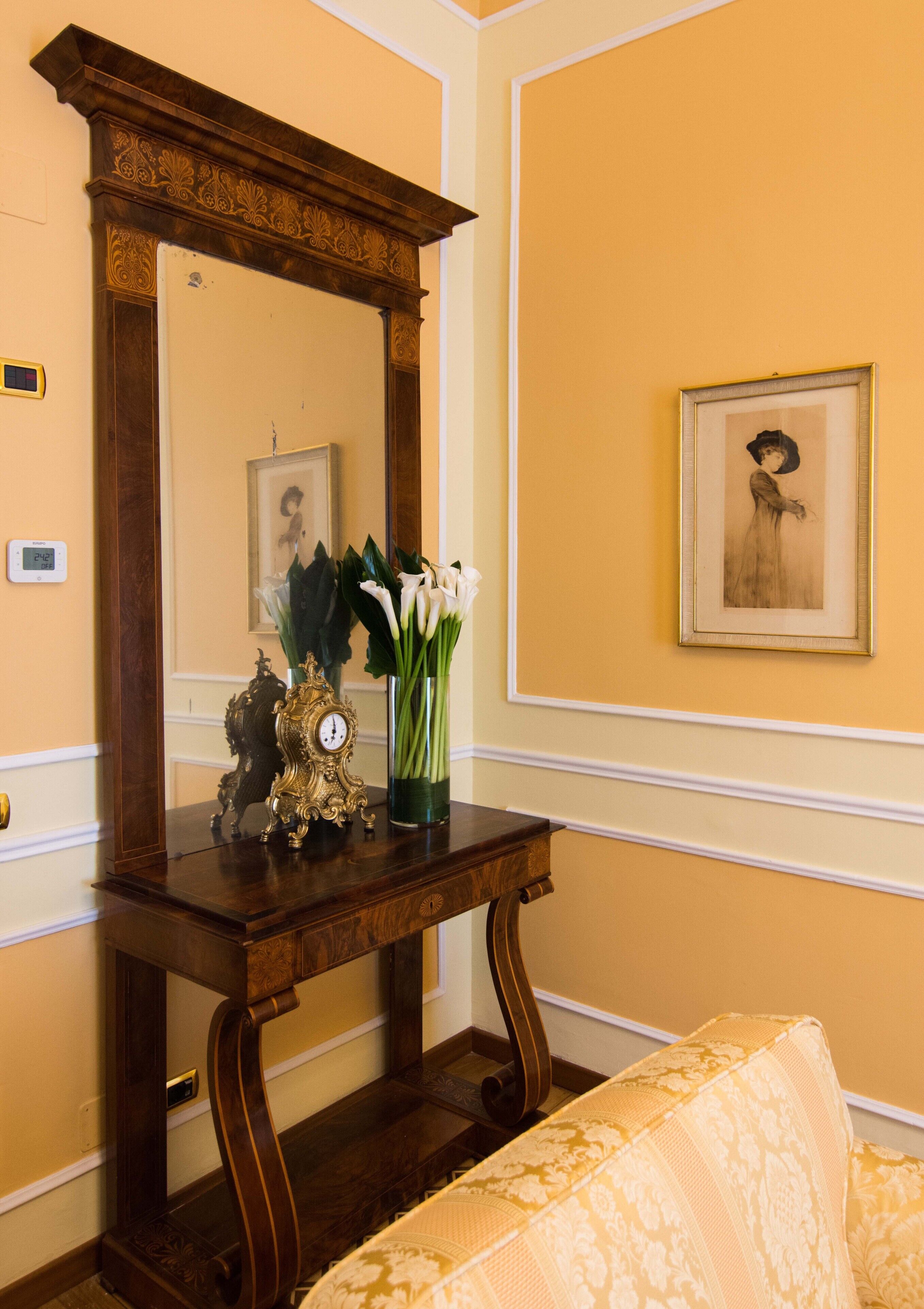 Una consola de madera con un reloj y flores, un cuadro en la pared y un sofá estampado.