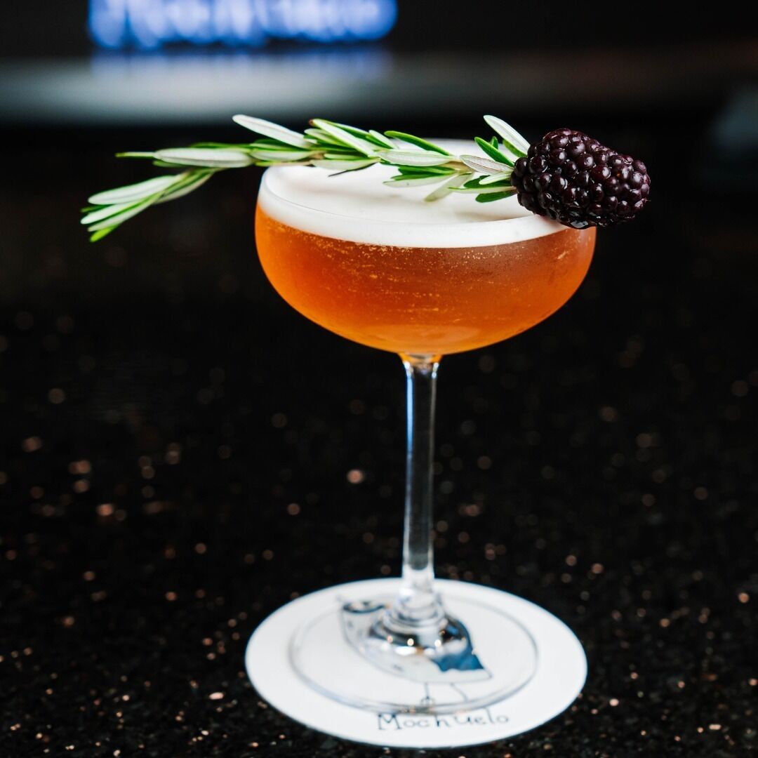 En cocktail i et stilkskank med en bjørnebærpynt og rosmarinkvist.