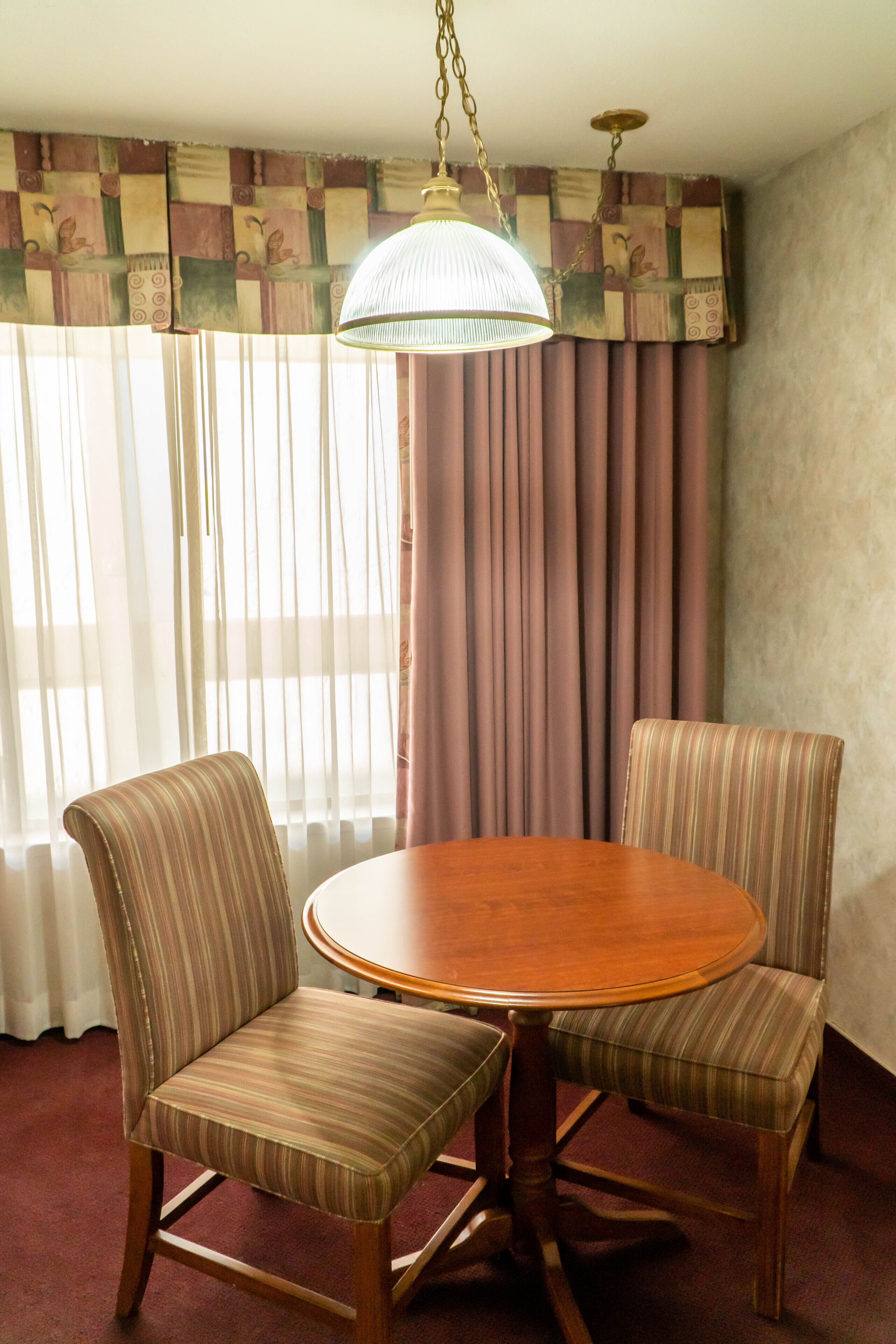 A dining area with a round wooden table and two striped chairs, a hanging lamp, and a window with curtains.