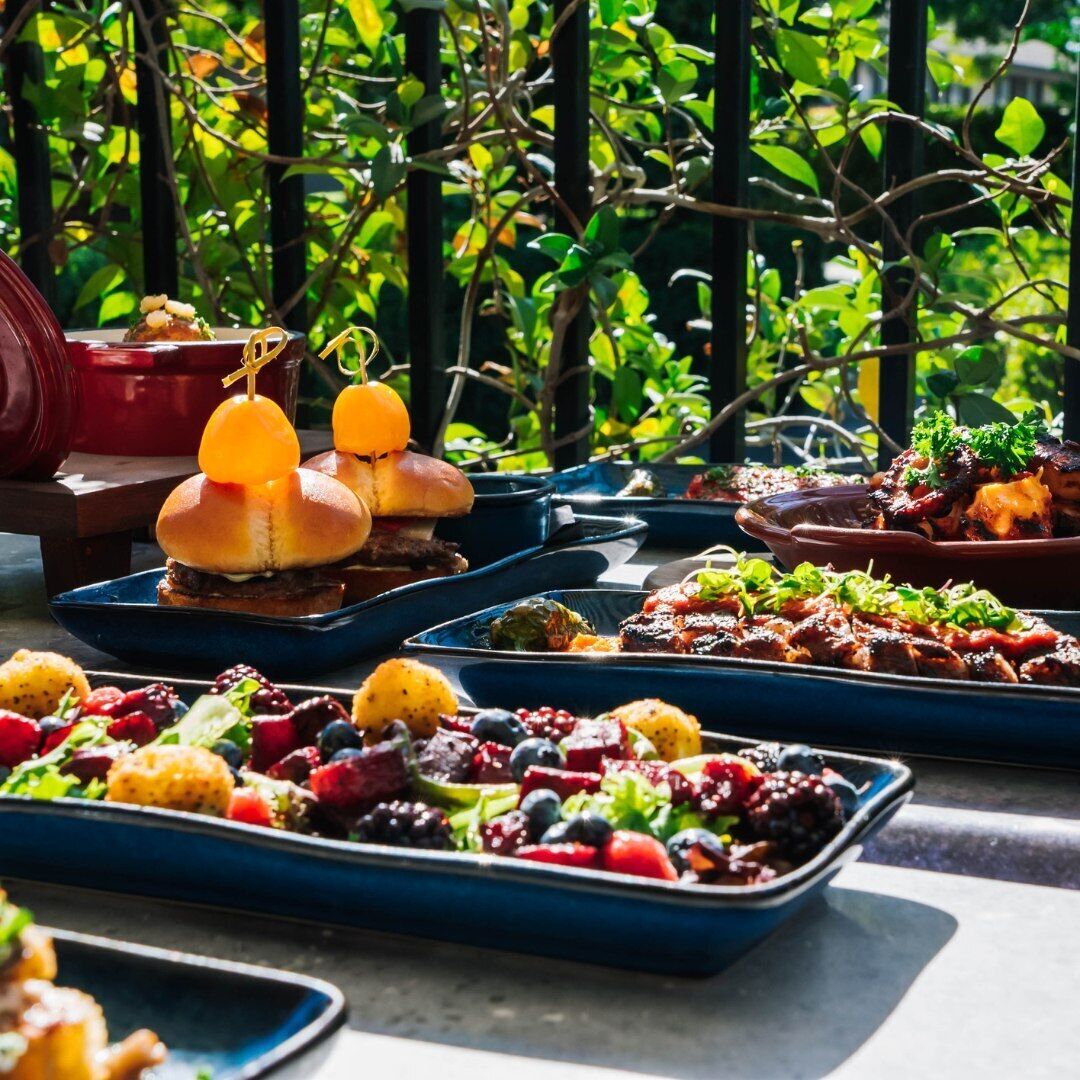 A variety of dishes on a table, including burgers, grilled meats, and a fruit salad.