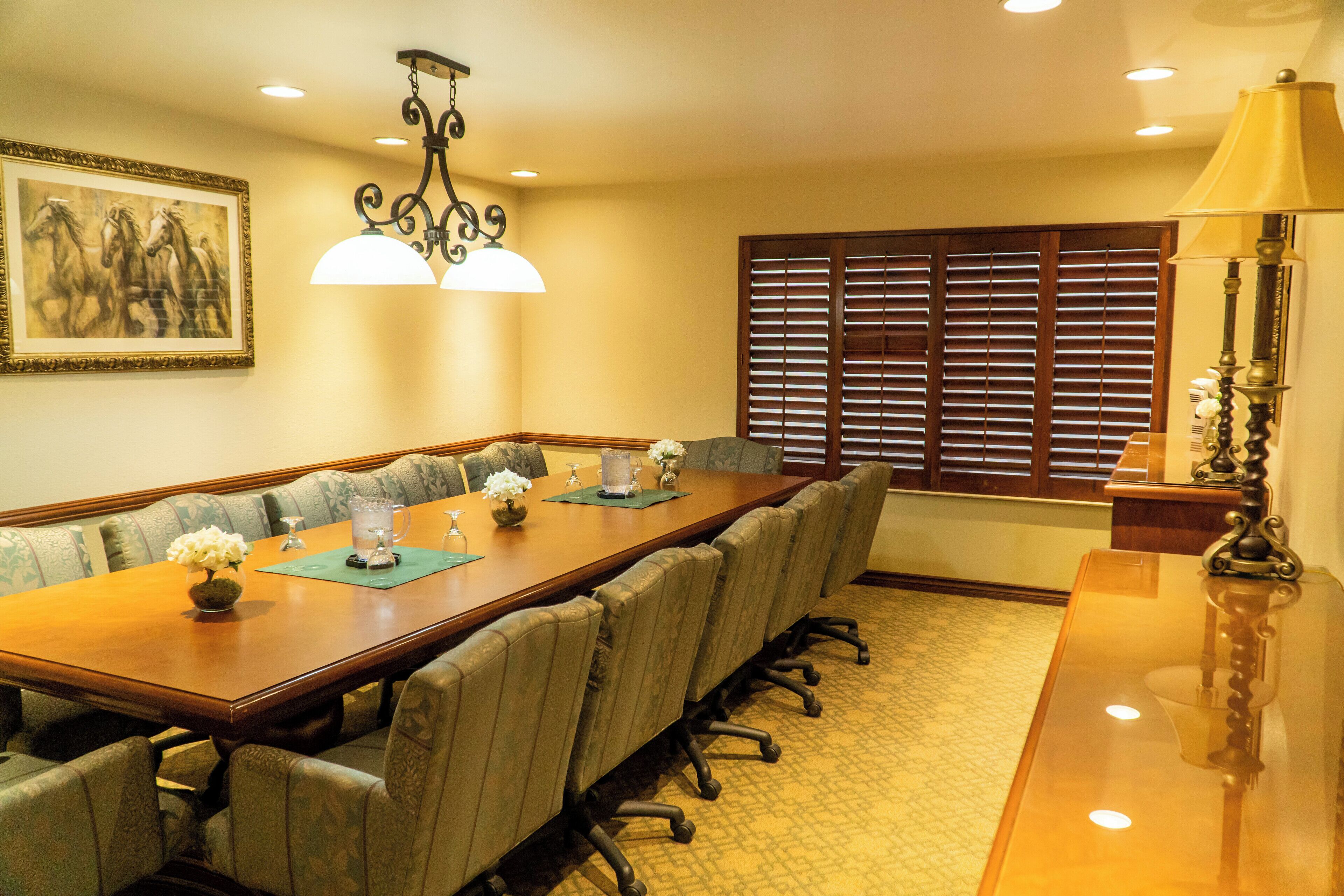 A conference room with a long table, chairs, a chandelier, a window with blinds, and a lamp.