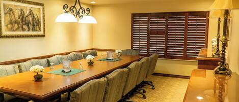 A conference room with a long table, chairs, a chandelier, a window with blinds, and a lamp.