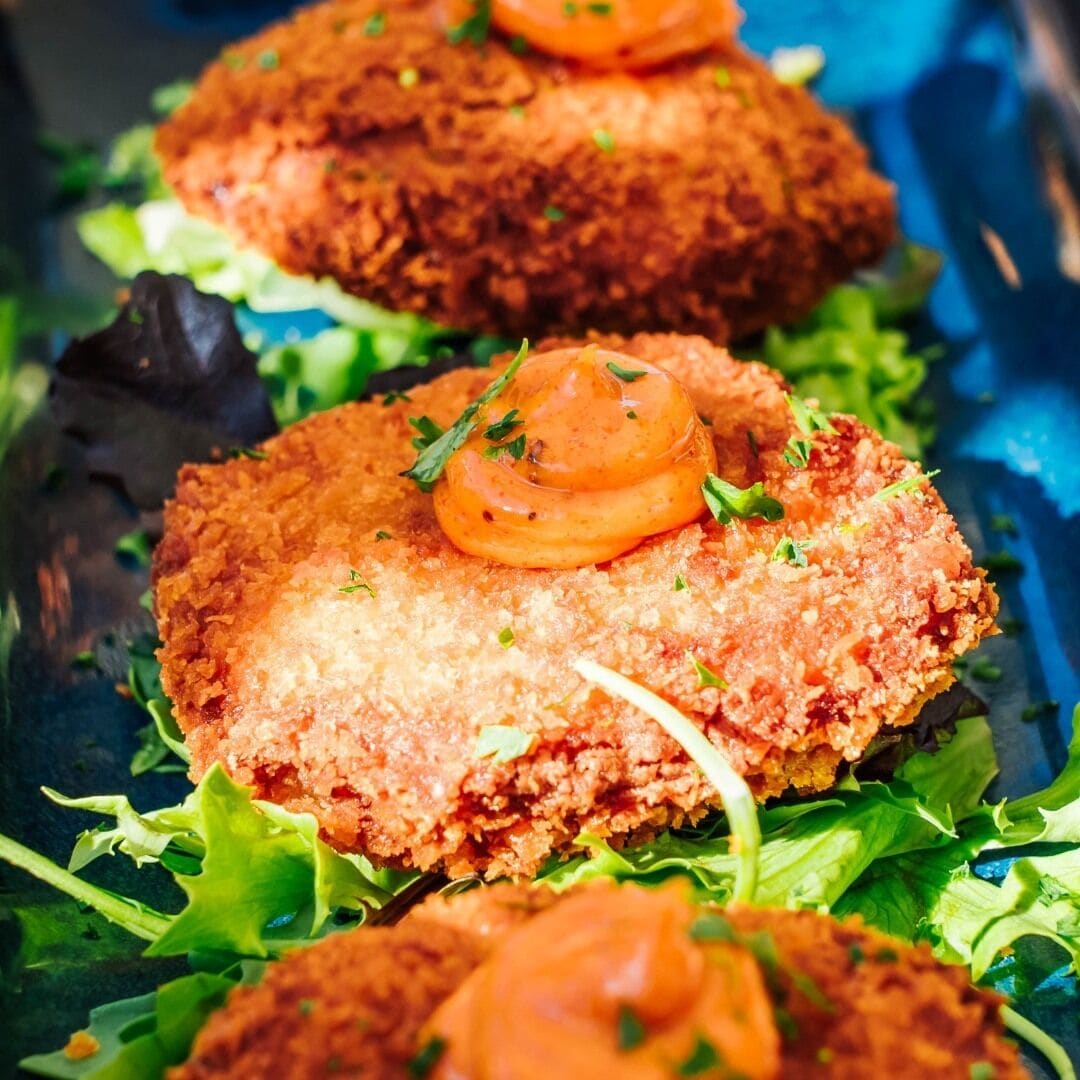 Gros plan sur un plat, vraisemblablement une galette panée et frite, garnie d’une tranche de tomate et d’herbes, et servie sur un lit de verdure.