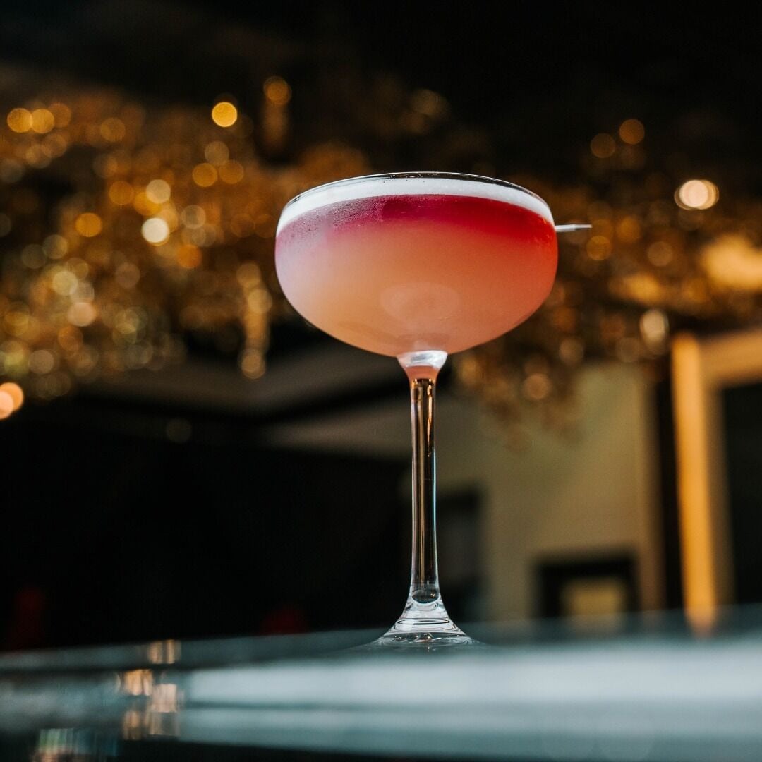 Een cocktail in een martini-glas met een kleurovergang, voor een onscherpe achtergrond met warme verlichting.