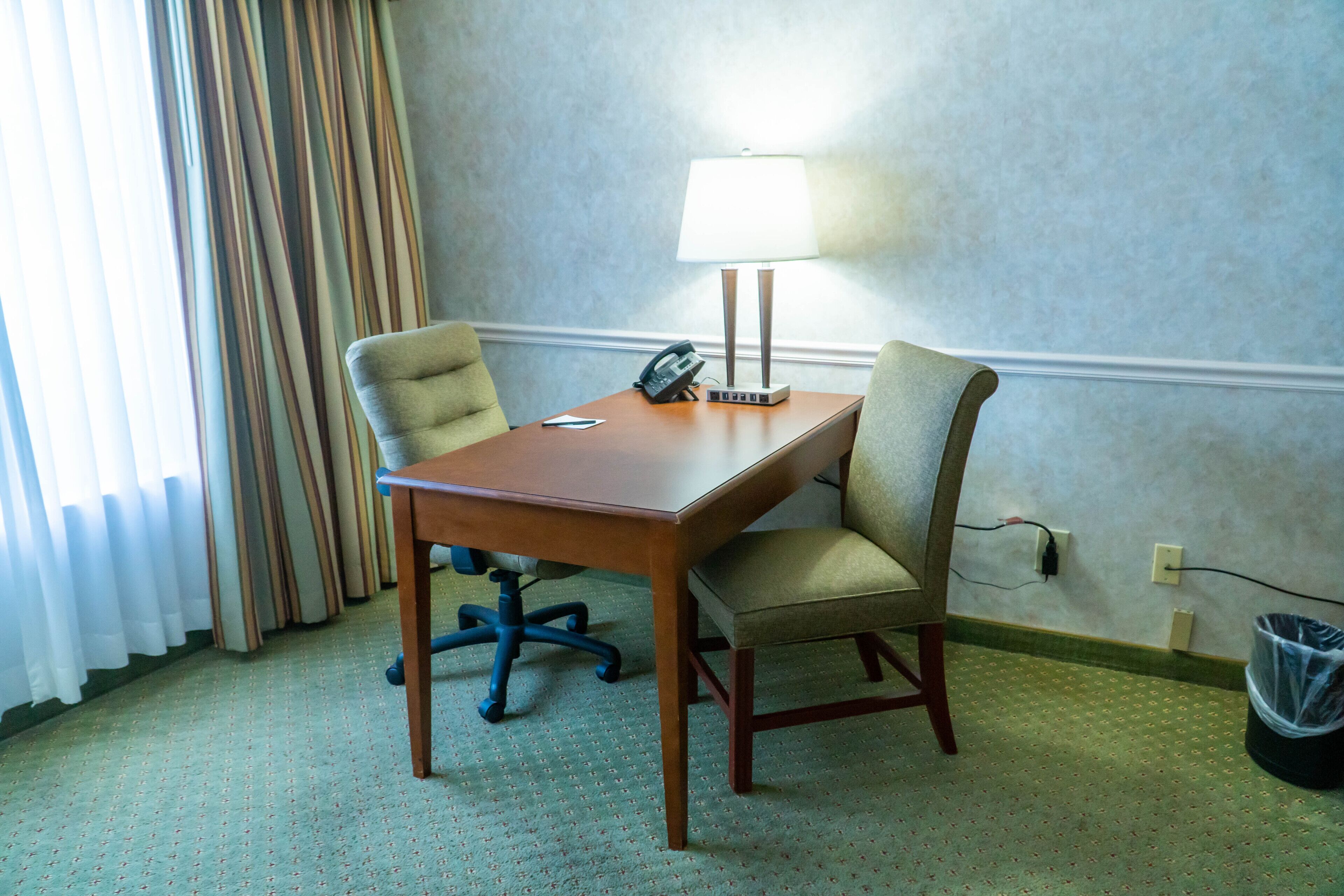 A hotel room with a wooden desk, two chairs, a lamp, a telephone, and a trash bin.