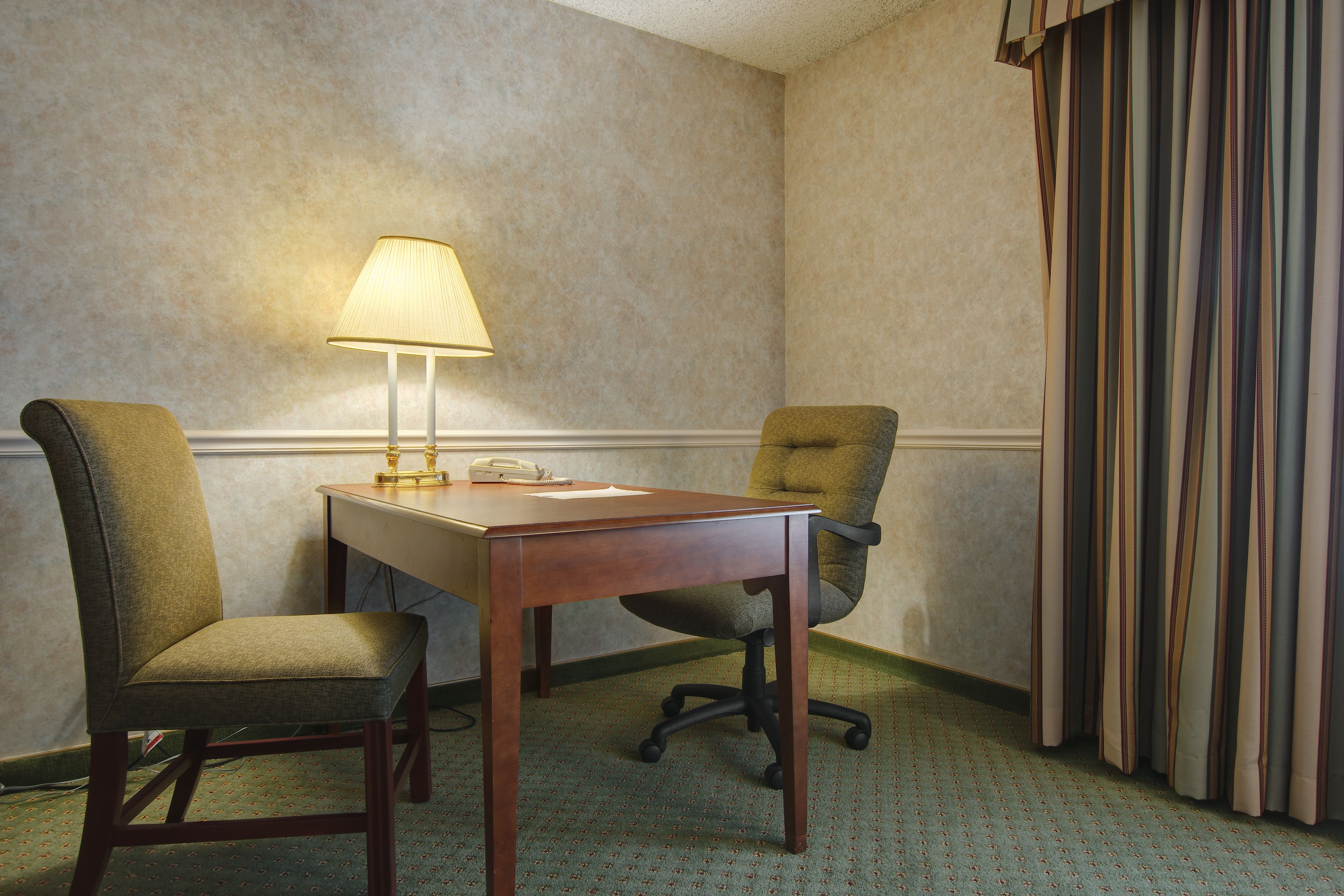 Habitación de hotel con escritorio de madera, dos sillas, una lámpara y una cortina a rayas.